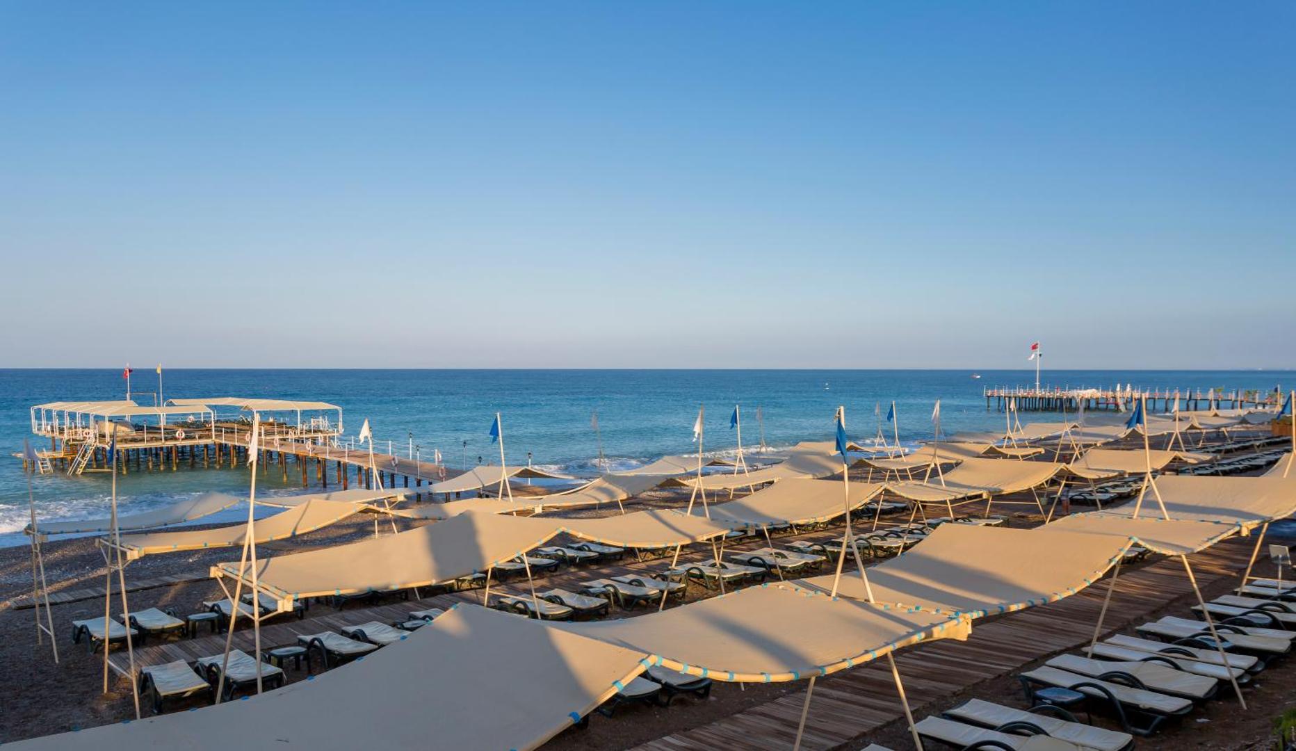 Mukarnas Spa & Resort plašā oļu pludmale ar daudziem sauļošanās krēsliem zem nojumēm un koka piestātni Alānijas reģionā