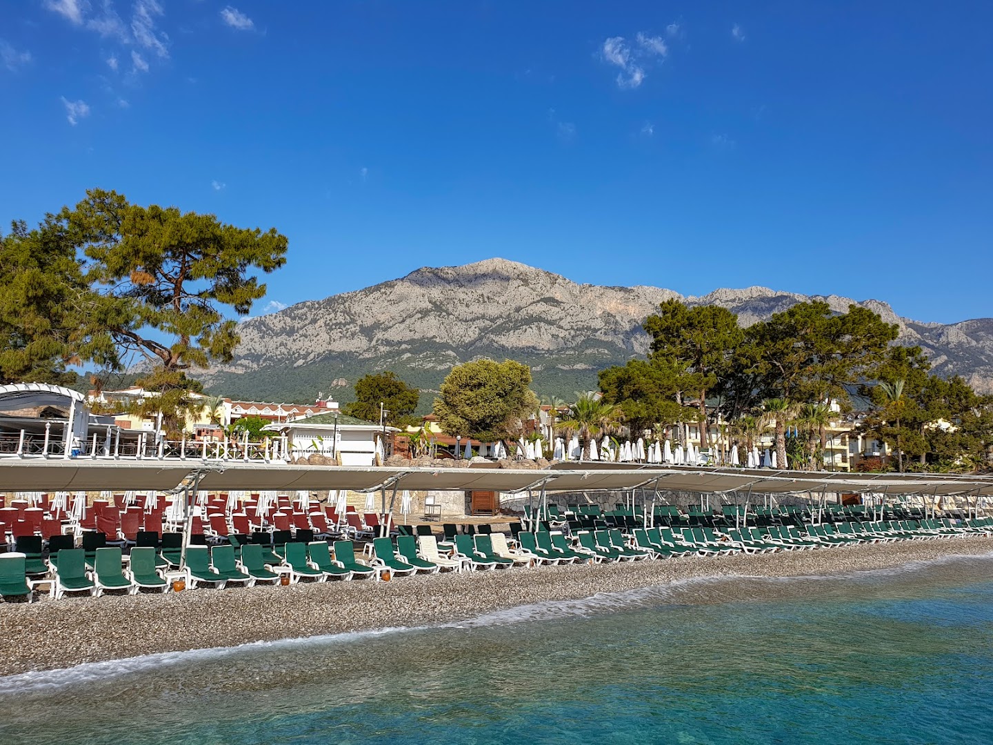Crystal Aura Beach Resort & Spa oļu pludmale ar sauļošanās krēsliem zem nojumēm un skatu uz kalniem Kemeras reģionā