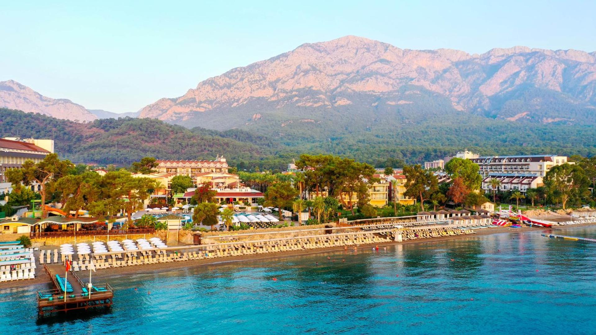 Crystal Aura Beach Resort & Spa privātā pludmale ar saulessargiem un gultām uz iespaidīgu kalnu fona Kemerā