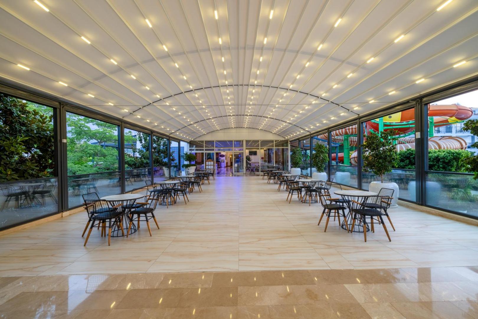 Crystal Aura Beach Resort & Spa moderna āra terase ar nojumi un galdiņiem — fonā redzami viesnīcas akvaparka ūdens slidkalniņi