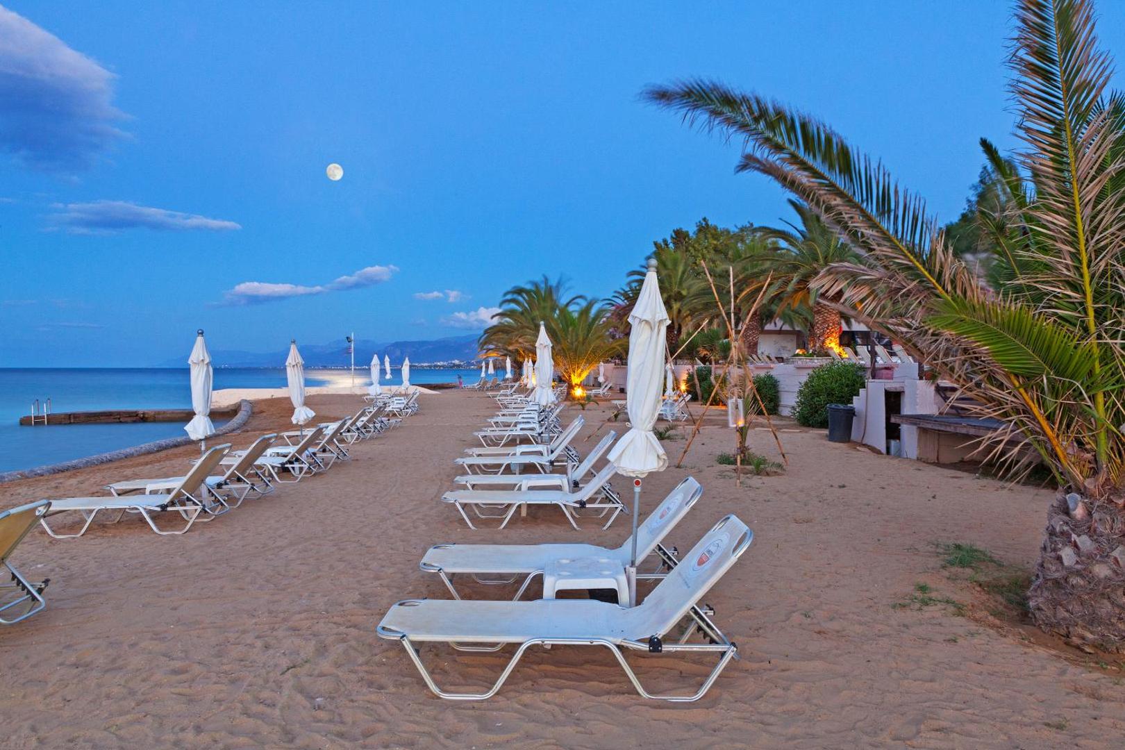Long Beach Resort privātā smilšu pludmale krēslas stundā — sauļošanās krēsli, saulessargi un palmas pie mierīgas jūras Krētā