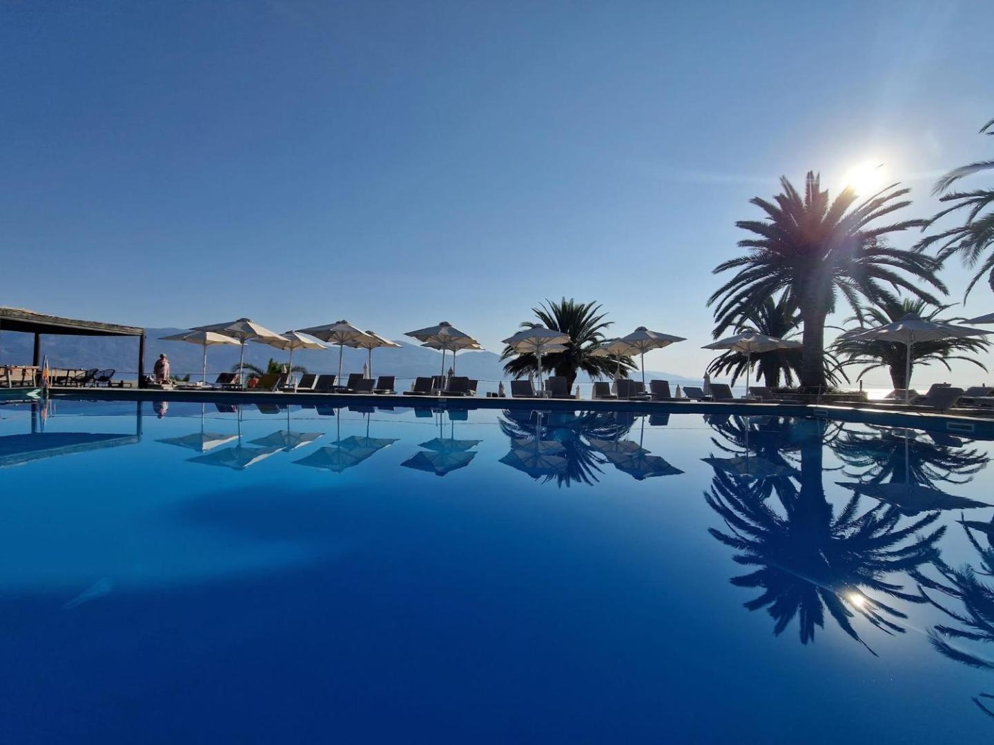 Skats uz Long Beach Resort baseinu, kurā atspīd palmas un saulessargi, fonā redzami kalni un jūra saules gaismā.