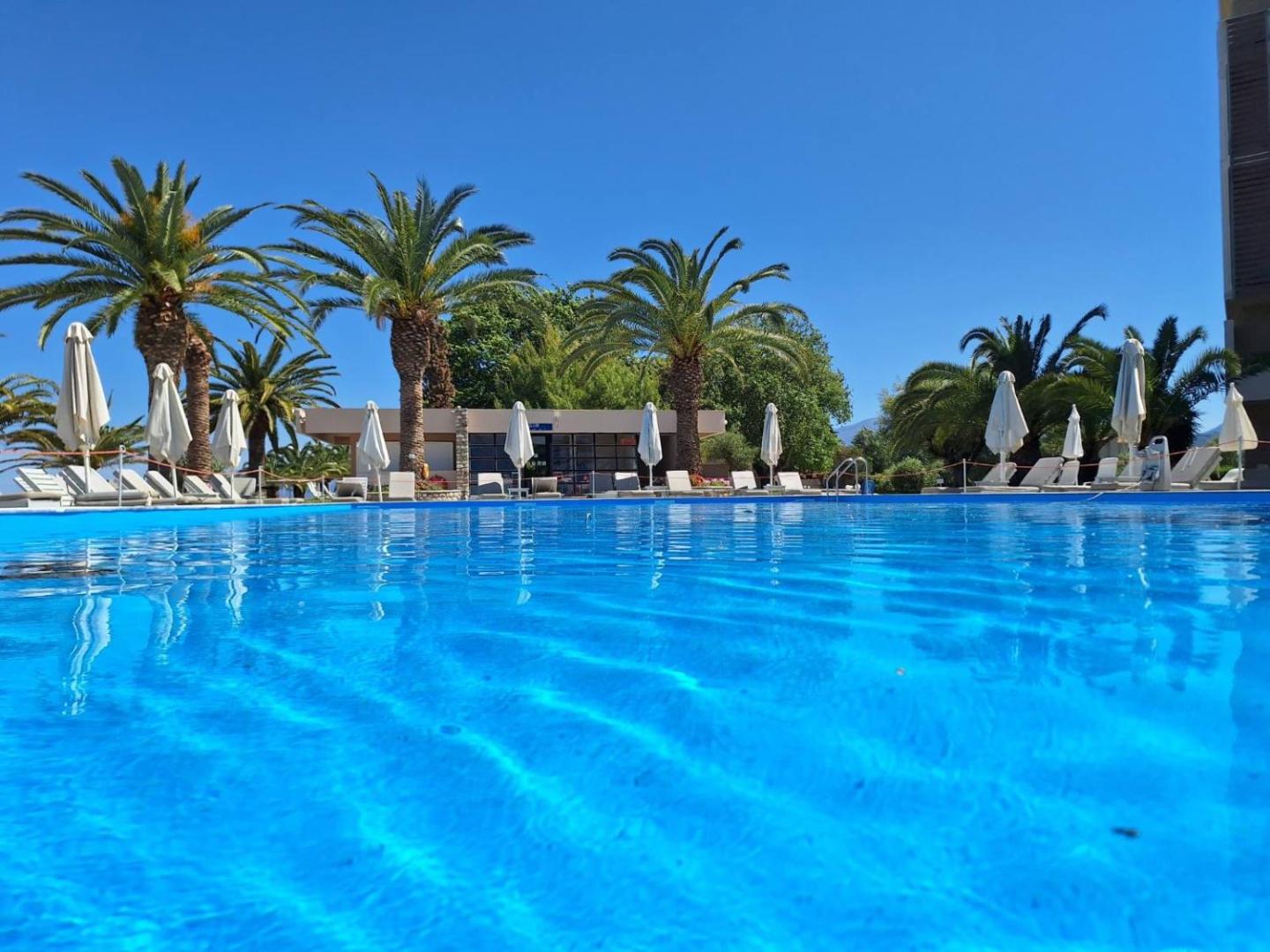 Long Beach Resort āra baseins ar zilu ūdeni, ko ieskauj palmas un balti saulessargi ar sauļošanās krēsliem saulainā dienā.