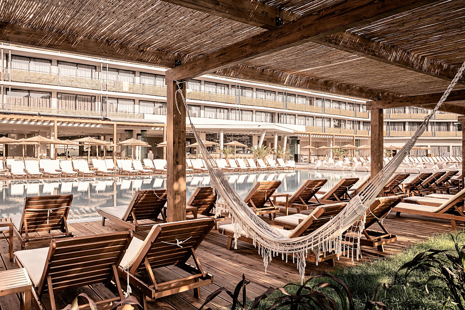 Sunny Beach Club atpūtas zona pie baseina ar koka zviļņiem, šūpuļtīklu un saulessargiem zem koka nojumes Saulainajā Krastā.