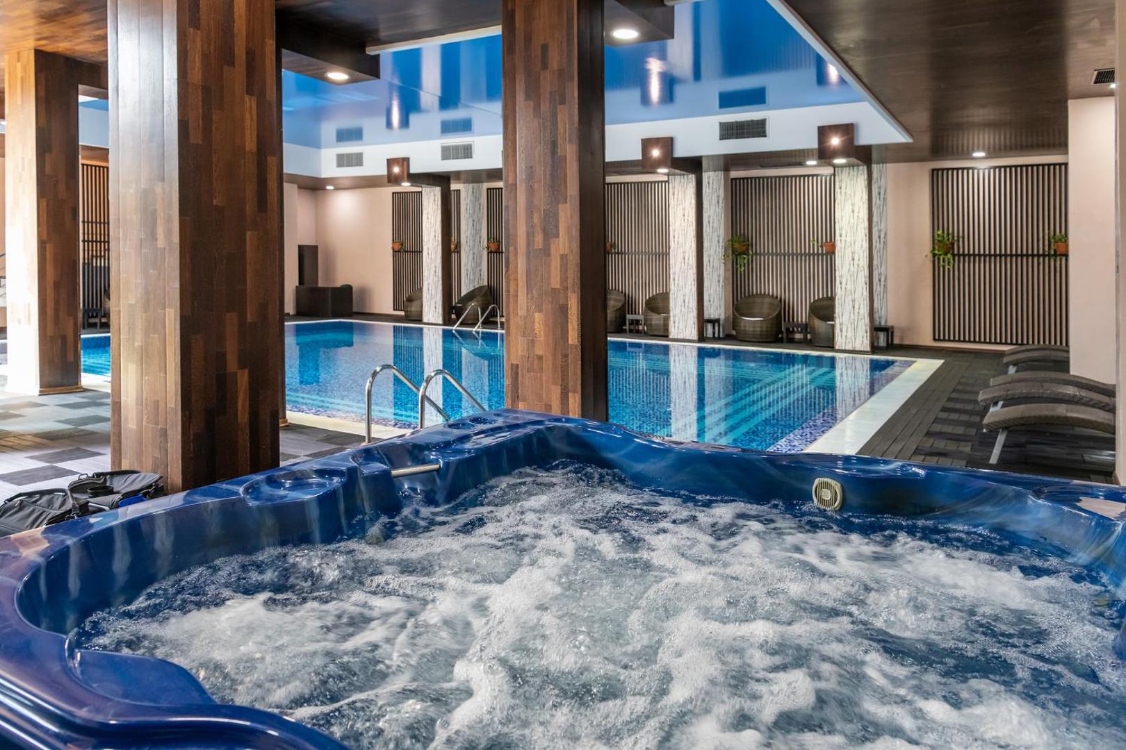 Garden Of Eden spa zona ar burbuļvannu priekšplānā un lielu iekštelpu baseinu fonā, koka kolonnu apdare