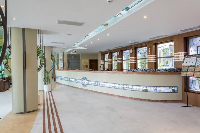 Garden Of Eden viesnīcas recepcijas zona ar garu reģistrācijas leti, vitrāžām un gaišu, plašu vestibilu
