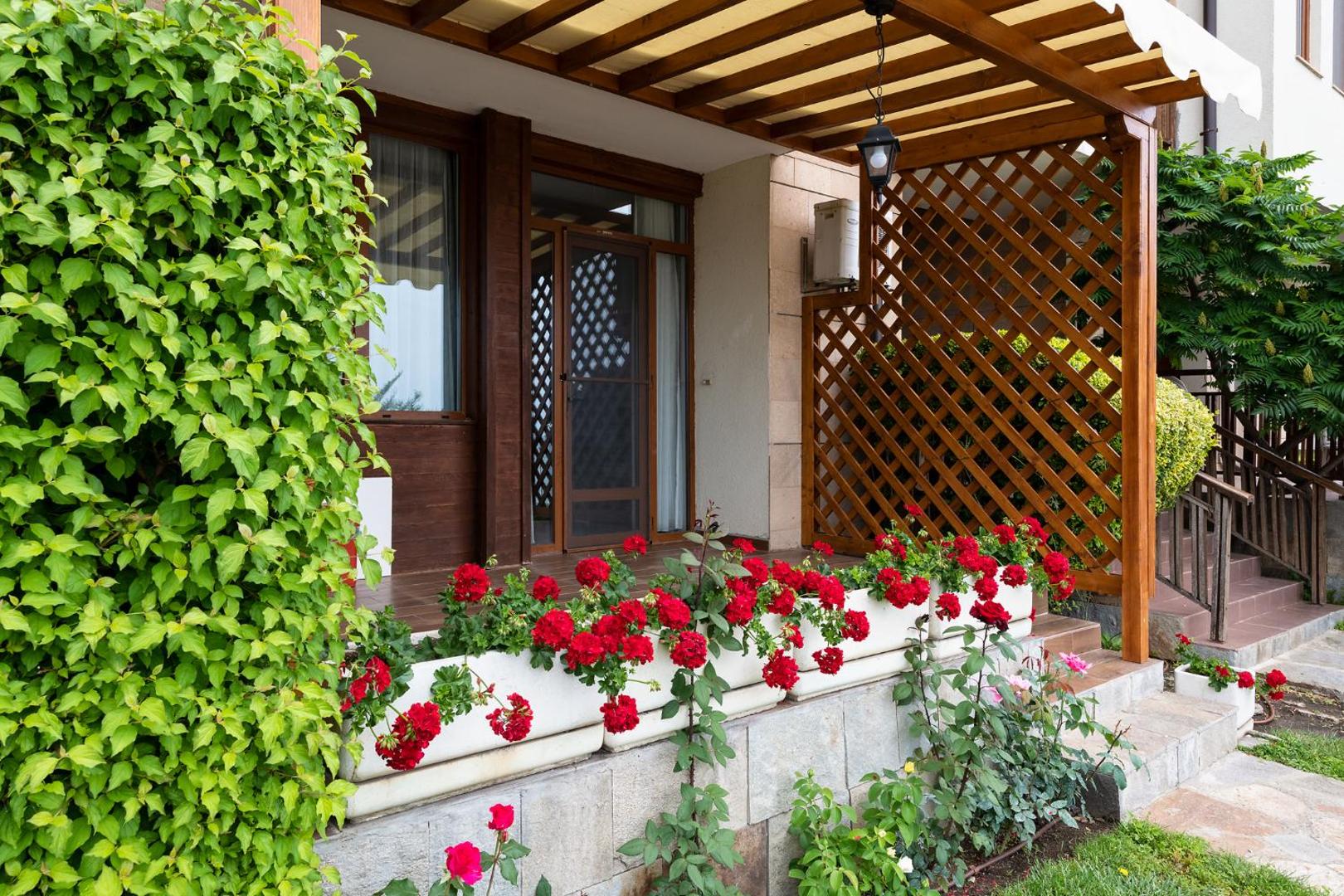Garden Of Eden viesnīcas numura privātā terase ar koka pergolu, zaļumiem un košām sarkanām pelargonijām podos