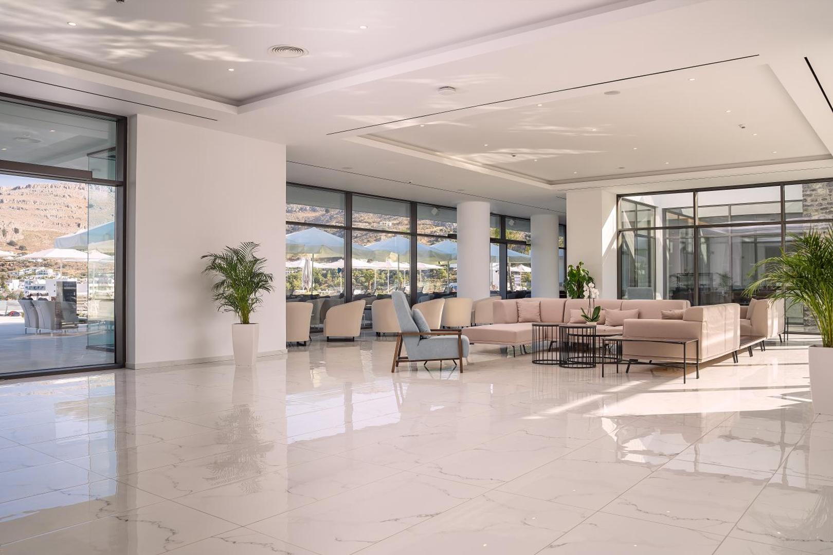 Lindos Grand Resort & Spa plašā un gaišā lobby atpūtas zona ar moderniem dīvāniem, marmora grīdu un skatu uz Rodas piekrasti
