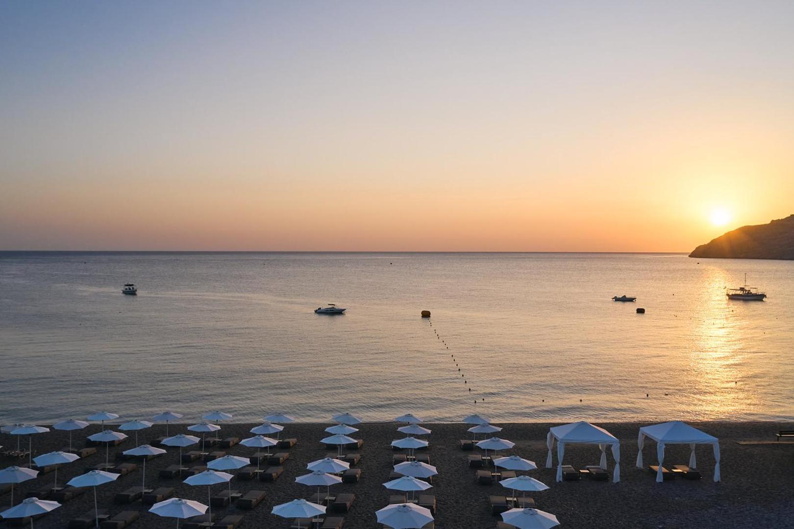 Lindos Grand Resort & Spa privātā pludmale saulrieta laikā ar saulessargiem un atpūtas teltīm Rodas piekrastē, skats uz mierīgu jūru