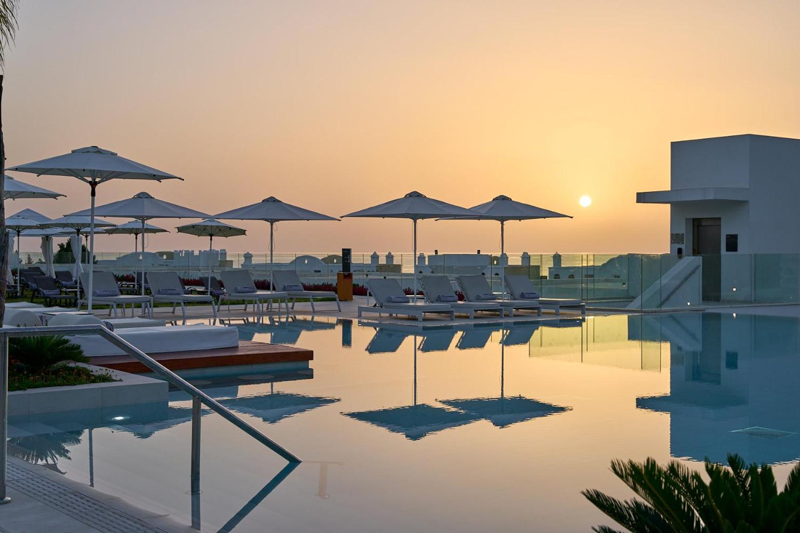 Lindos Grand Resort & Spa atpūtas zona pie baseina ar baltiem saulessargiem un ērtiem zviļņiem saulrieta fonā