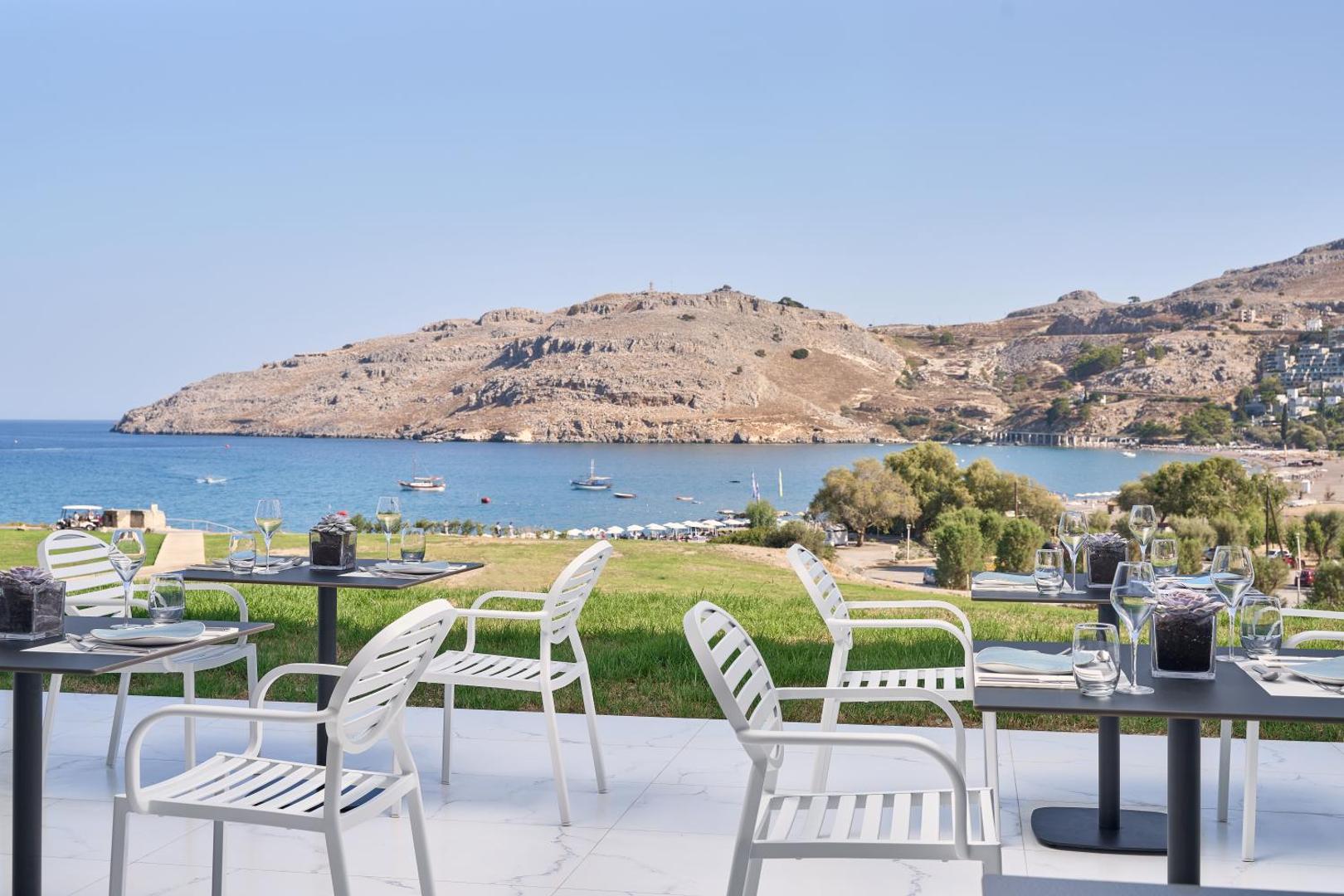 Lindos Grand Resort & Spa āra restorāna terase ar skatu uz jūru un kalniem Rodas salā, Grieķijā — balti krēsli un galdi