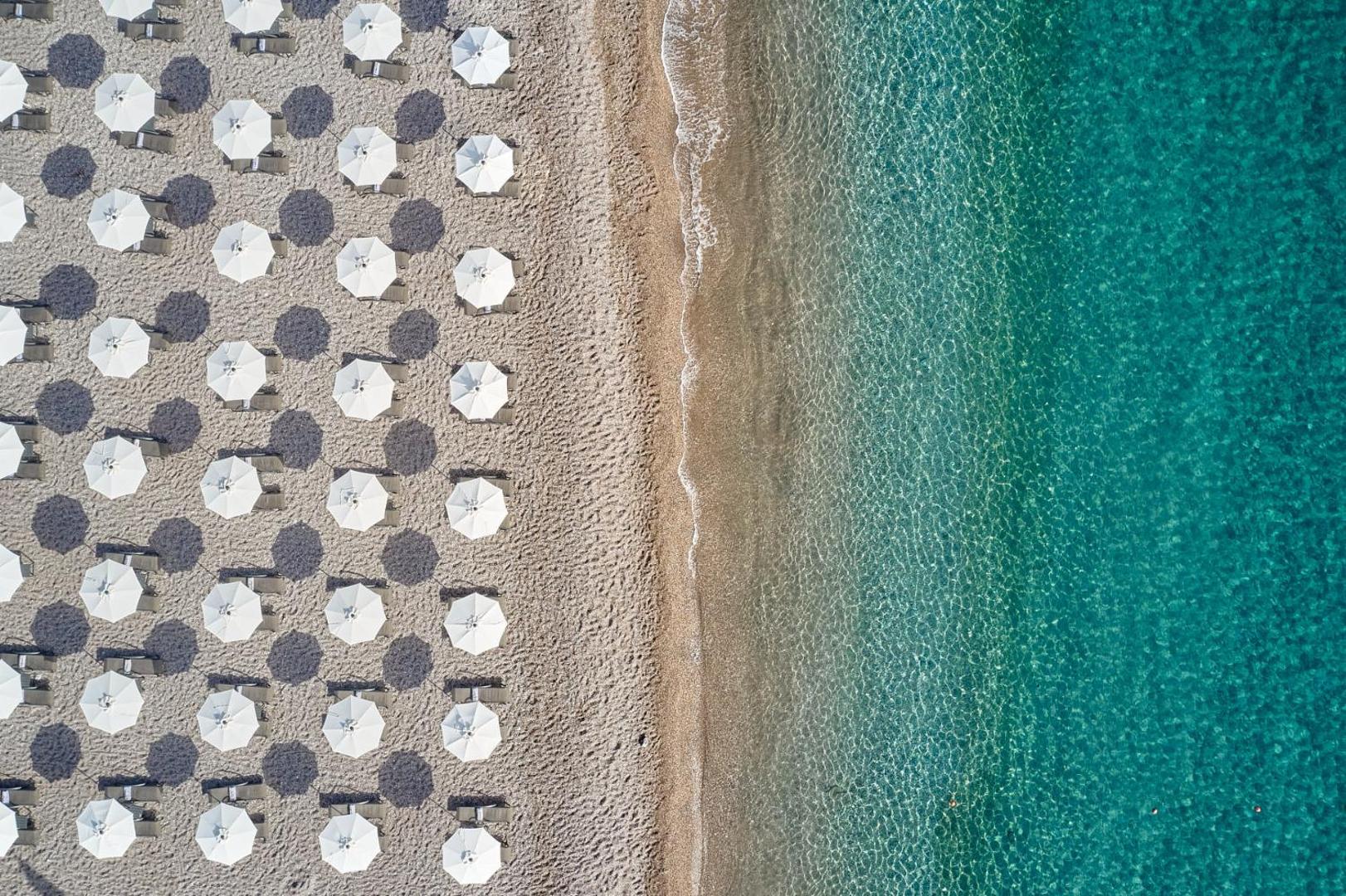 Lindos Grand Resort & Spa pludmales josla no augšas — balti saulessargi uz oļu pludmales fona un caurspīdīgs jūras ūdens Rodas salā