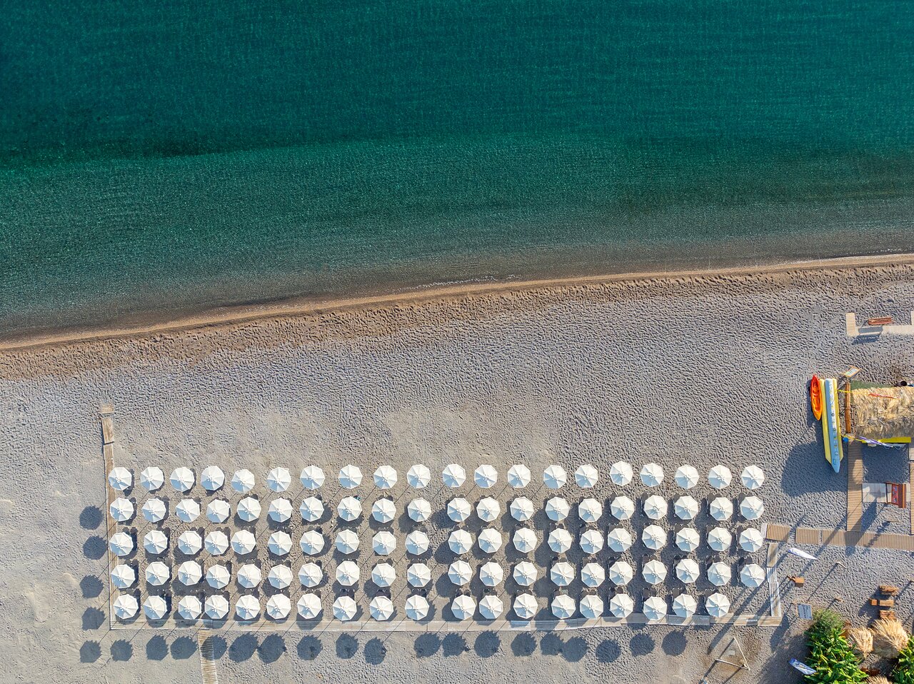 Lindos Grand Resort & Spa pludmales skats no putna lidojuma — balti saulessargi sakārtotās rindās pie dzidra, tirkīzzila ūdens Rodā