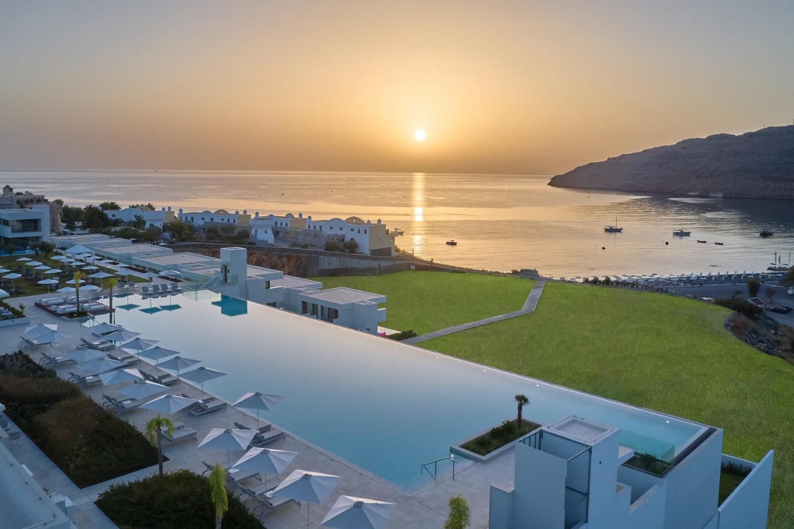 Lindos Grand Resort & Spa panorāmas skats no augšas uz baseinu, piekrasti un jūras līci Rodā saulrieta gaismā