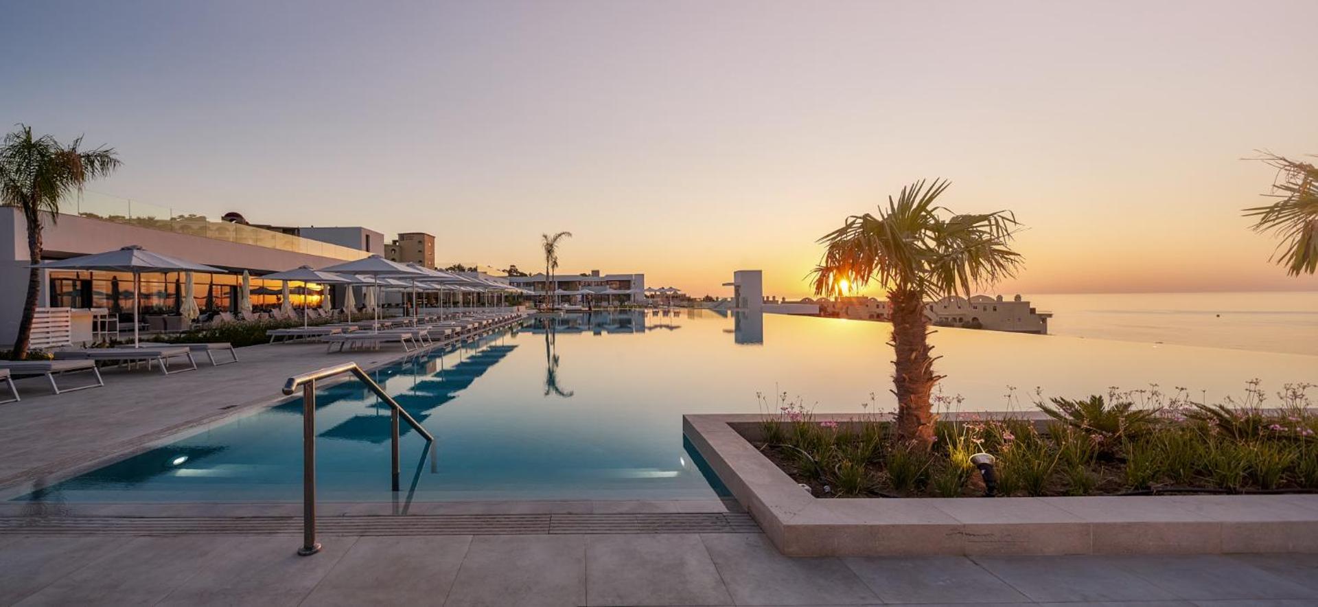 Lindos Grand Resort & Spa bezgalības baseins saulrieta laikā Rodas salā ar skatu uz jūru, sauļošanās krēsliem un palmām