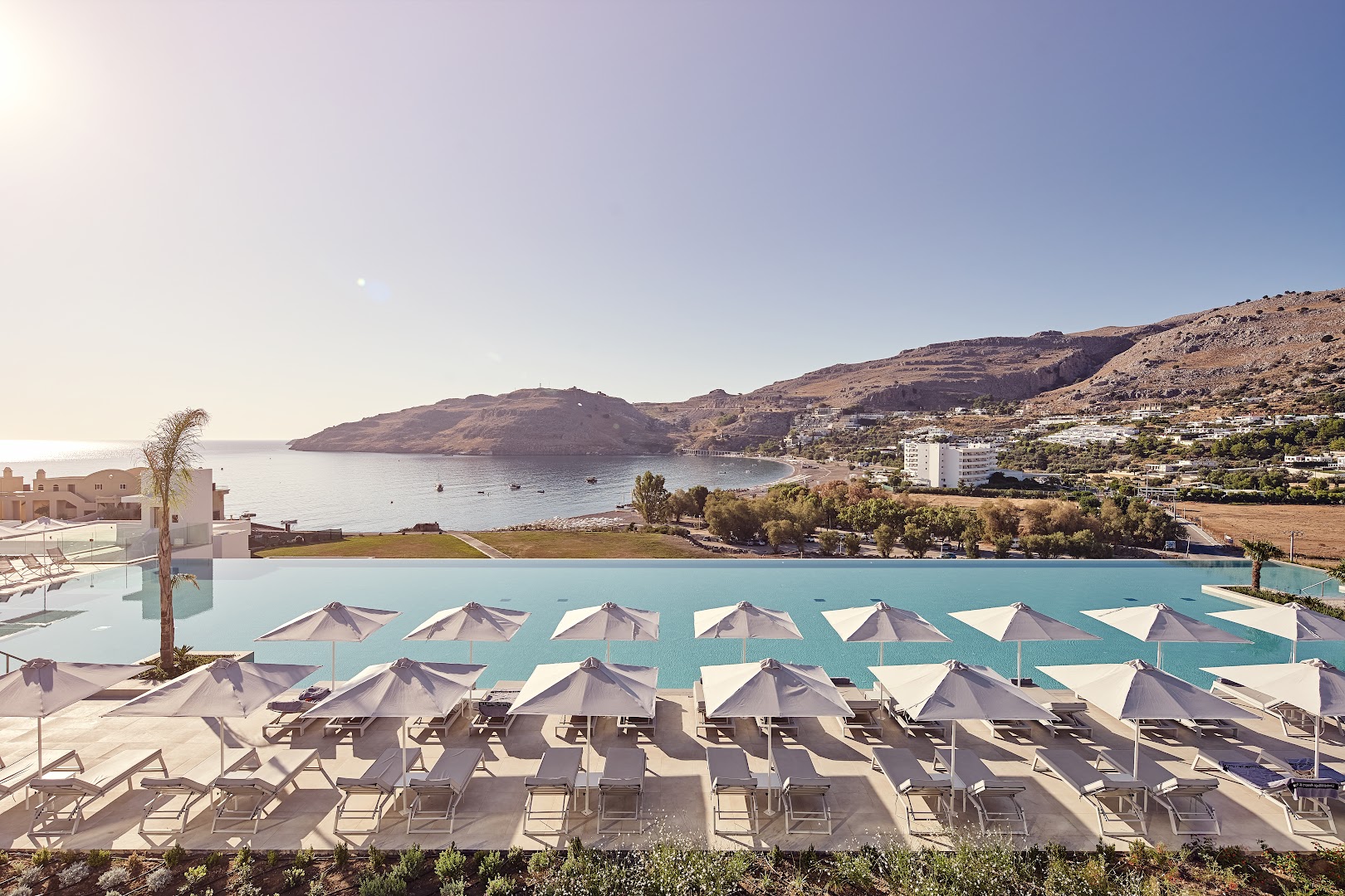 Lindos Grand Resort & Spa plašais baseins ar daudziem saulessargiem un skatu uz kalnaino Rodas piekrasti dienas laikā