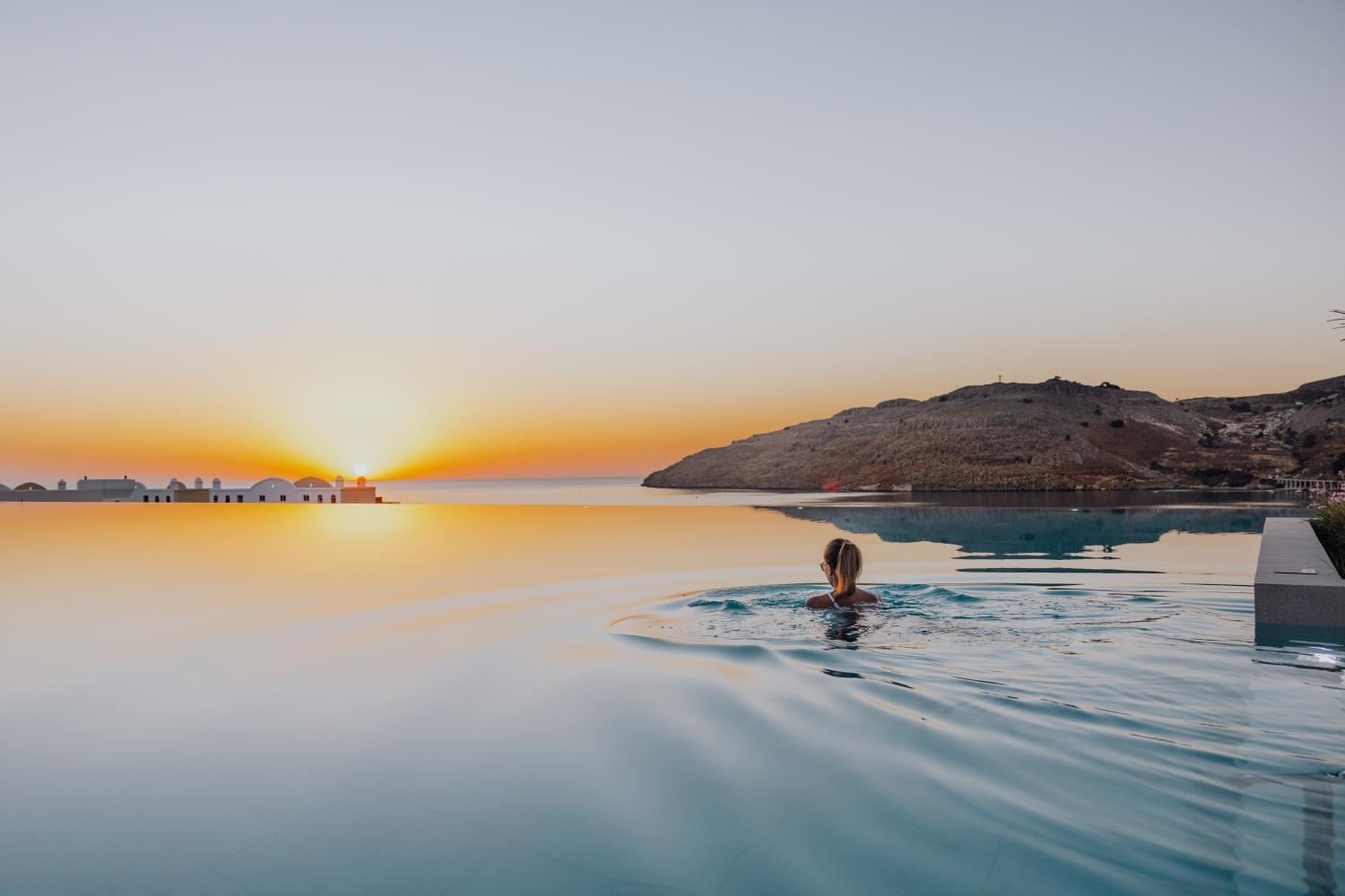 Sieviete peldas Lindos Grand Resort & Spa bezgalības baseinā saulrieta laikā ar skatu uz Rodas salas piekrastes klintīm