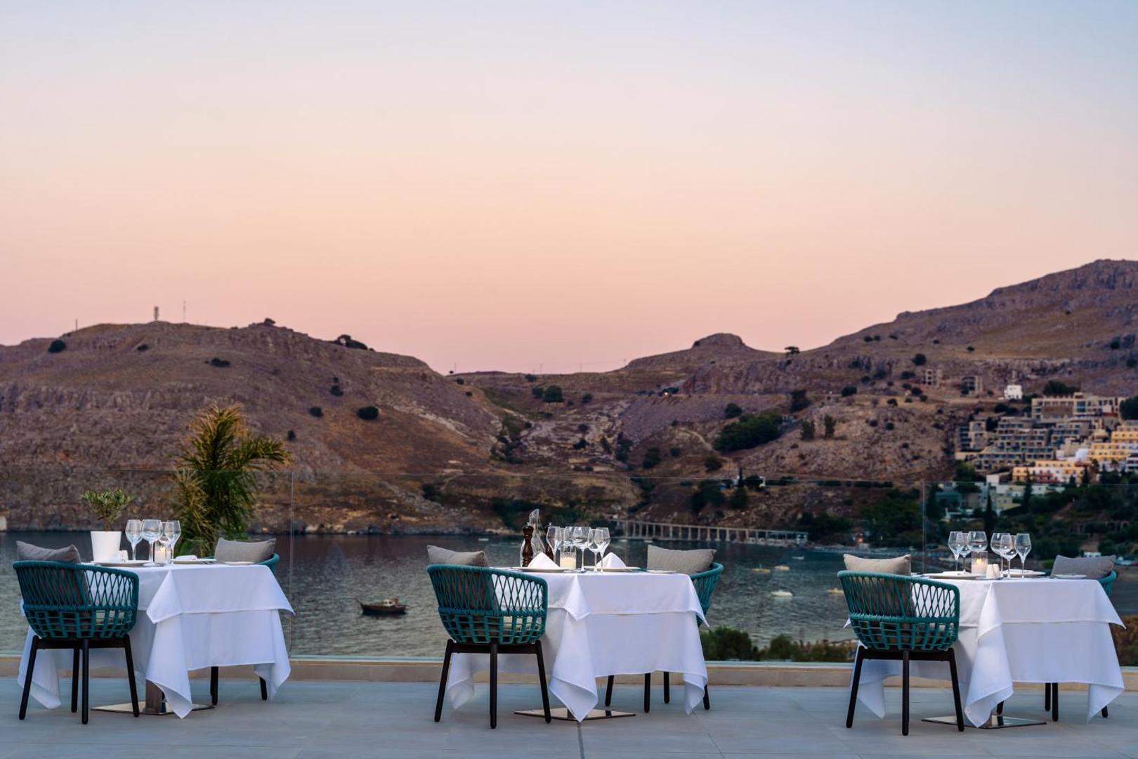 Lindos Grand Resort & Spa vakariņu galdi uz terases saulrieta laikā ar skatu uz līci un kalniem Rodas salā