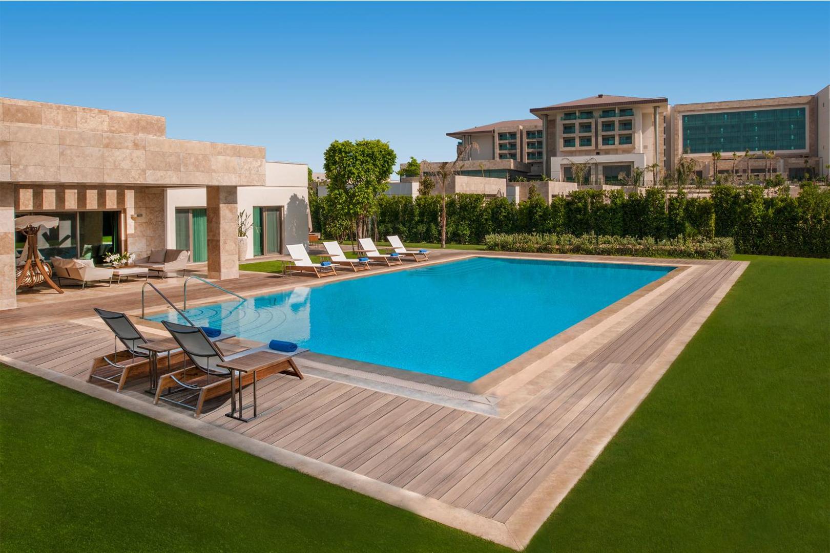 Regnum Carya villas āra baseins ar koka terasi un sauļošanās krēsliem dienas laikā — fonā redzama viesnīcas galvenā ēka