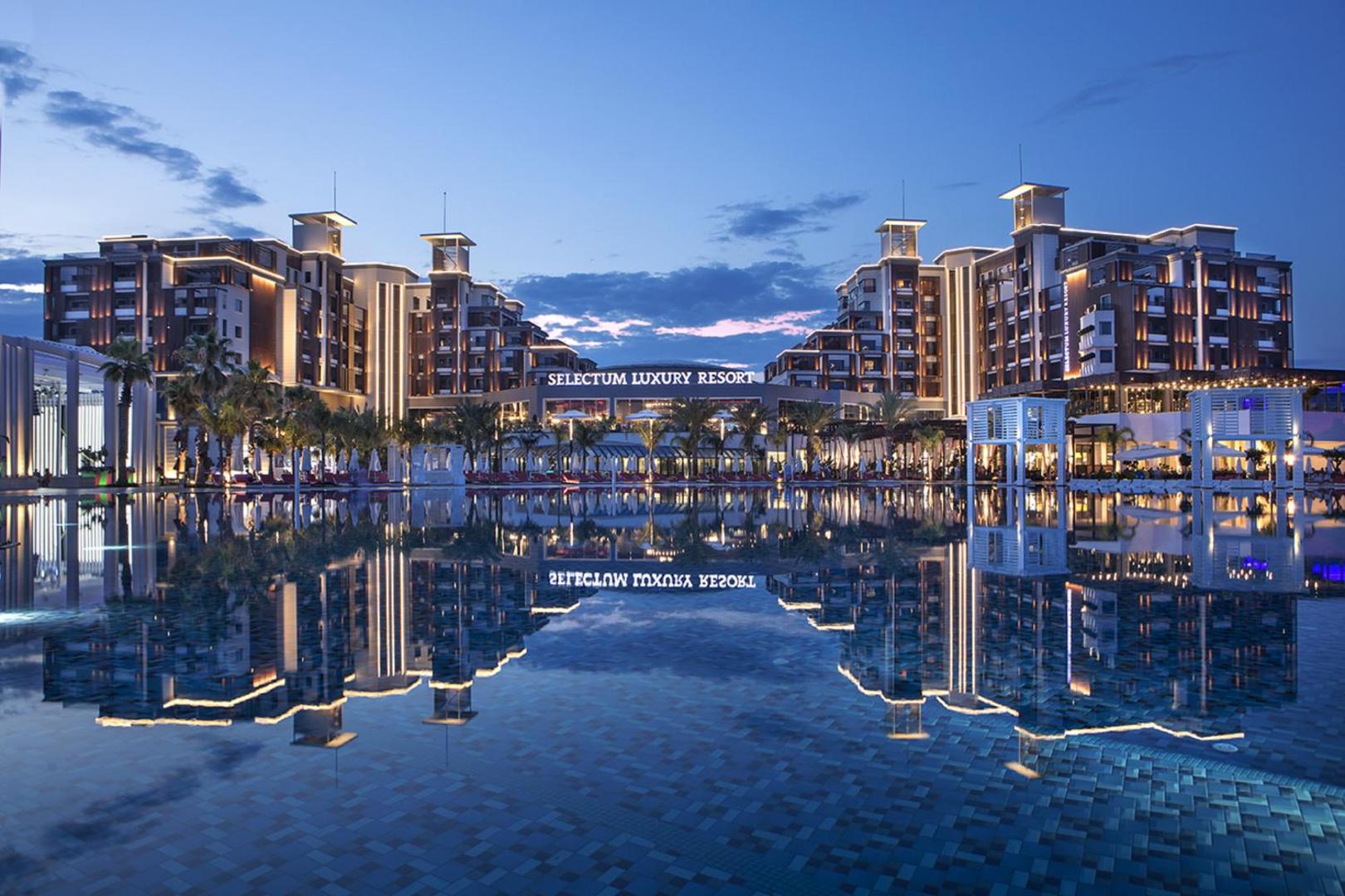Selectum Luxury Resort galvenā ēka vakarā ar apgaismojumu, kas atspīd lielajā āra baseinā Belekas kūrortā