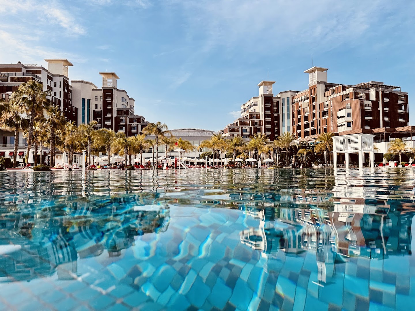 Selectum Luxury Resort galvenā ēka un palmas atspulgs dzidrā baseina ūdenī dienā — moderna arhitektūra un kūrorta vide