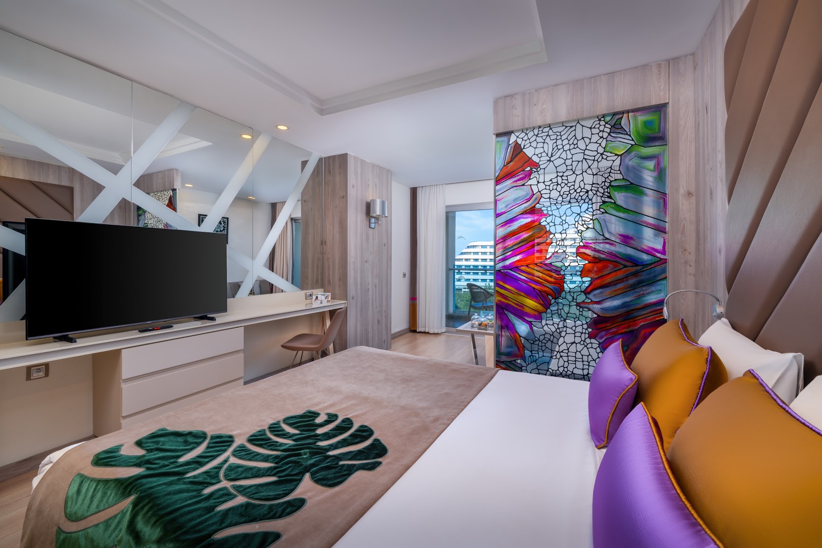 Delphin Be Grand Resort numura interjers ar lielu TV, dekoratīvu stikla sienu un skatu uz kūrorta teritoriju