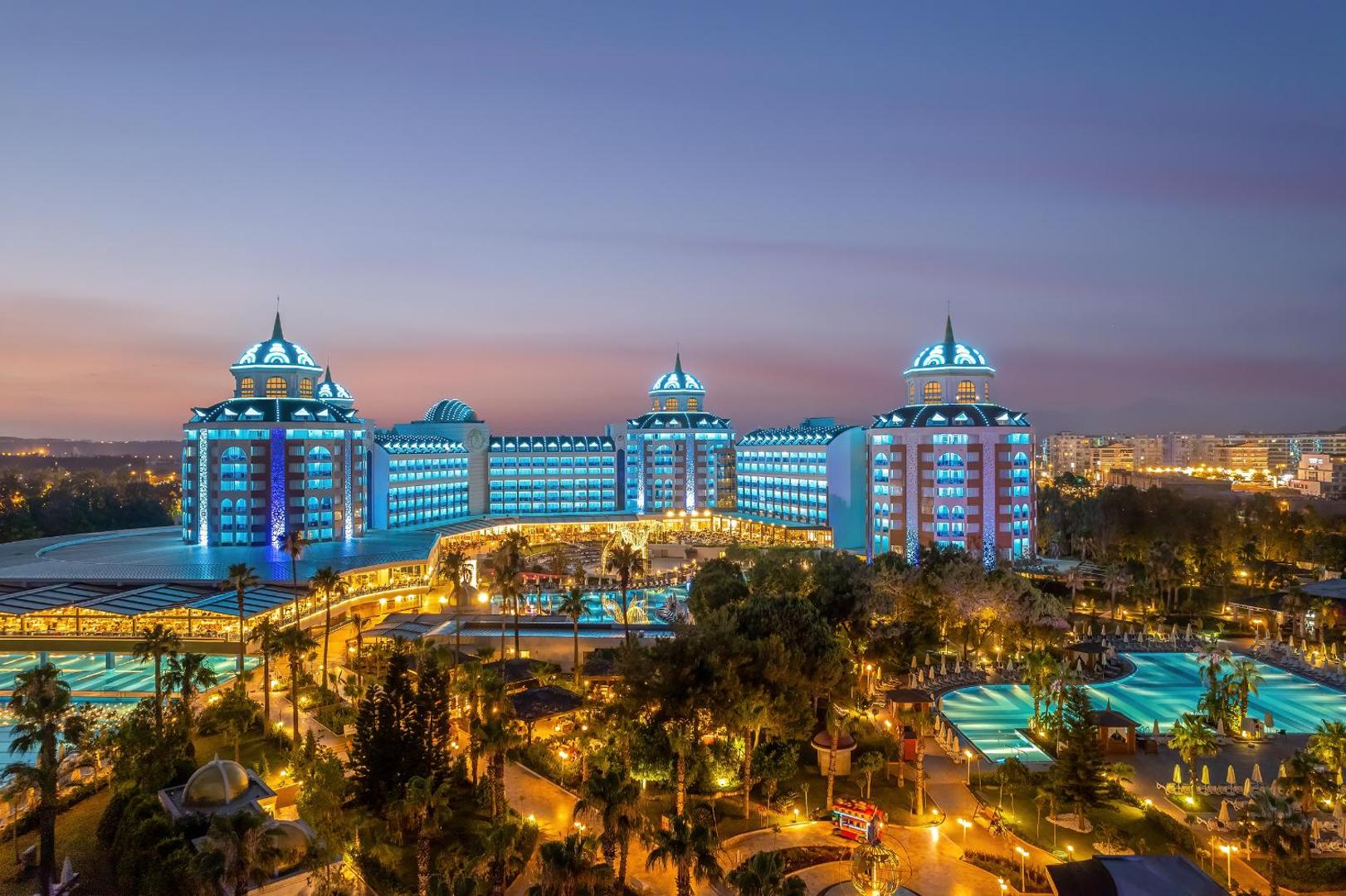 Delphin Be Grand Resort panorāmas skats vakarā — izgaismota viesnīcas fasāde, baseini un dārzs Antalijas piekrastē saulrieta laikā