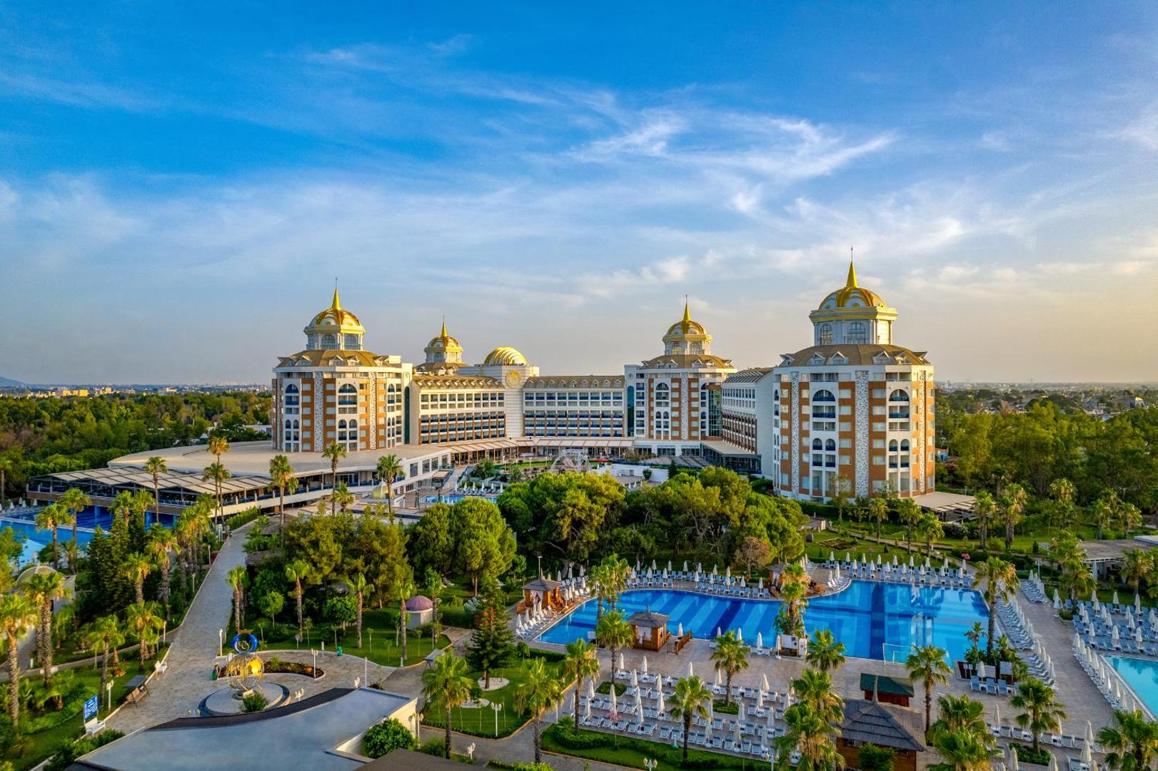 Delphin Be Grand Resort galvenā ēka un baseins saulrietā — palmas, sauļošanās krēsli un grezna arhitektūra Larā, Antalijā
