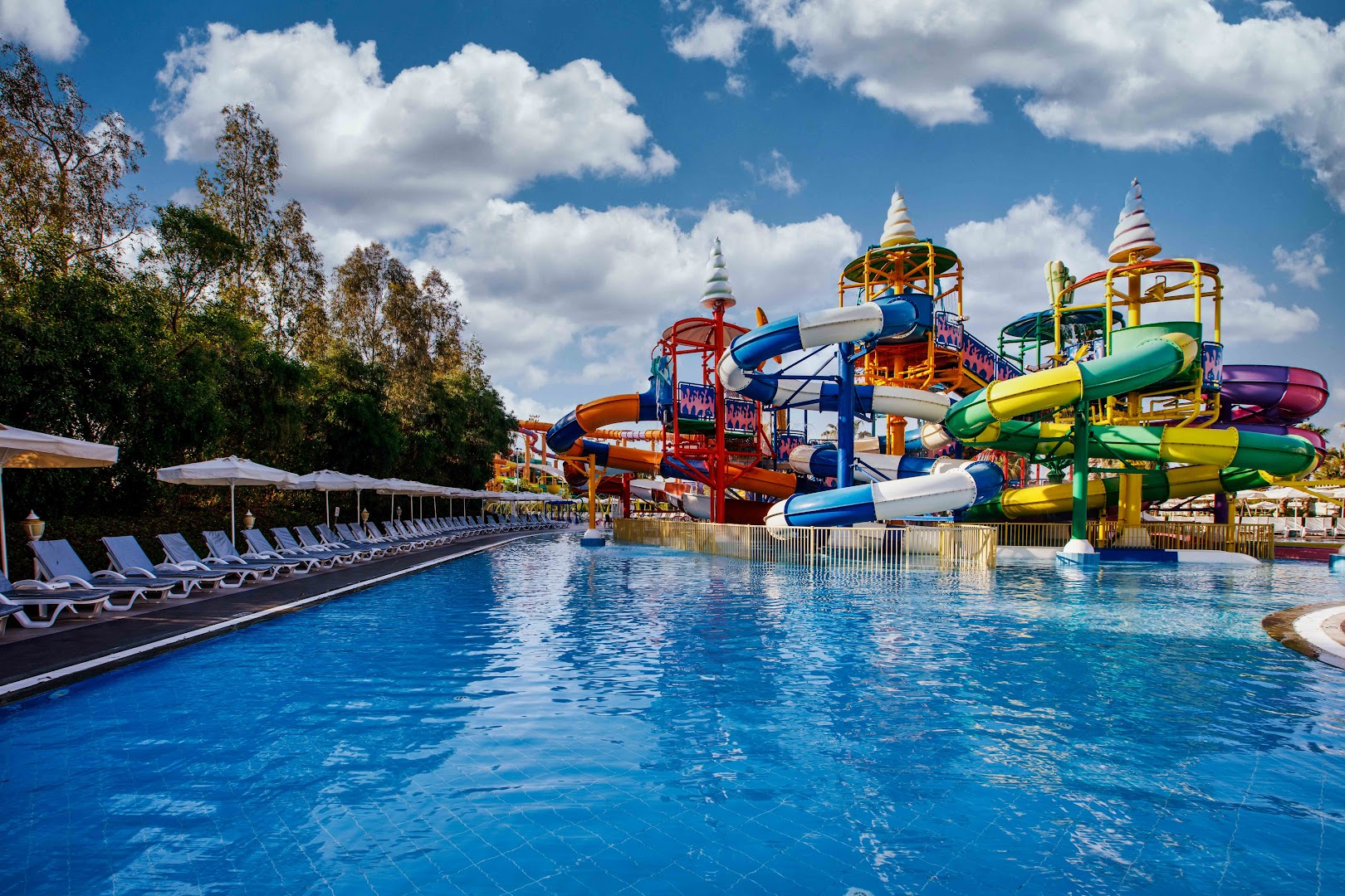 Delphin Be Grand Resort akvaparks ar krāsainiem ūdens slidkalniņiem un lielu āra baseinu Antalijā, Turcijā — sauļošanās zona ar saulessargiem
