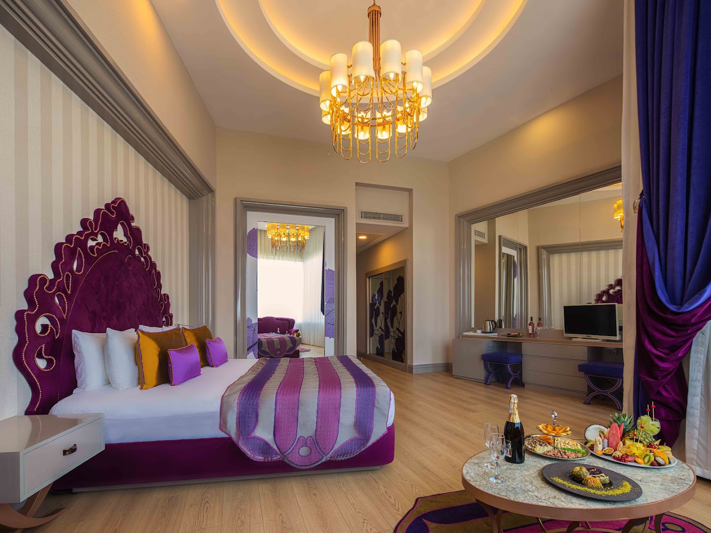 Delphin Be Grand Resort luksusa numurs ar violetiem akcentiem, greznu lustru un augļu plati uz galda