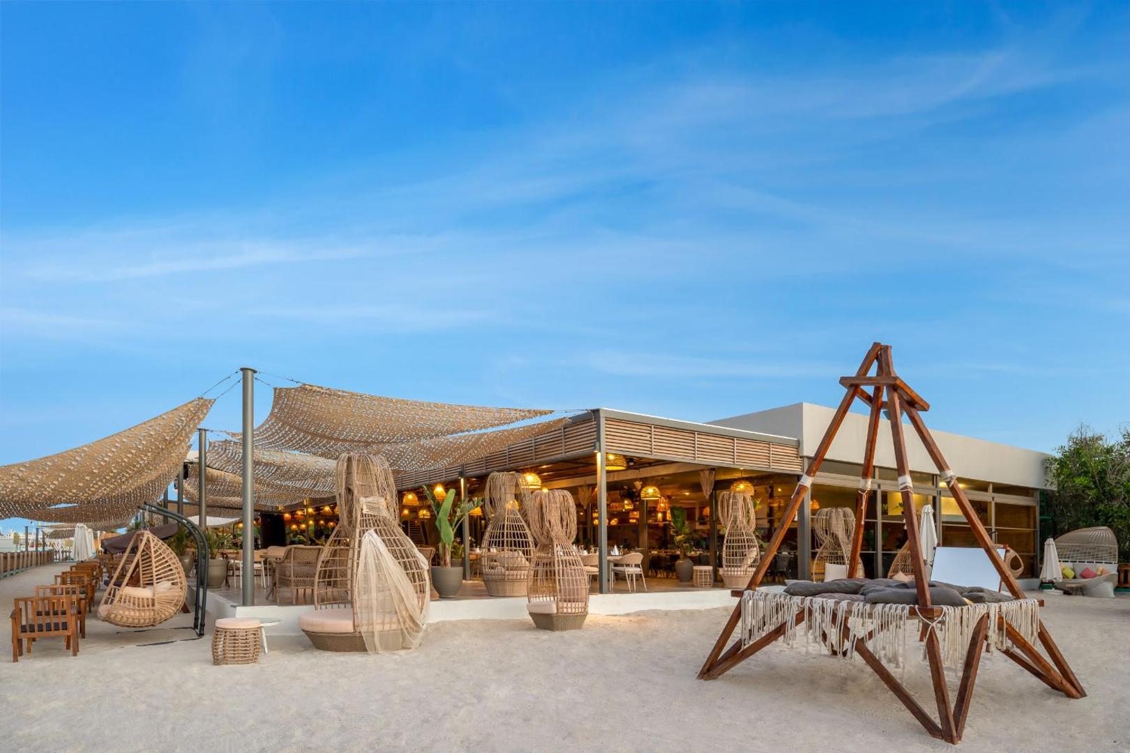 Delphin Be Grand Resort āra restorāns un bārs smilšainā pludmalē ar modernām nojumēm un pītiem dizaina krēsliem vakarā.