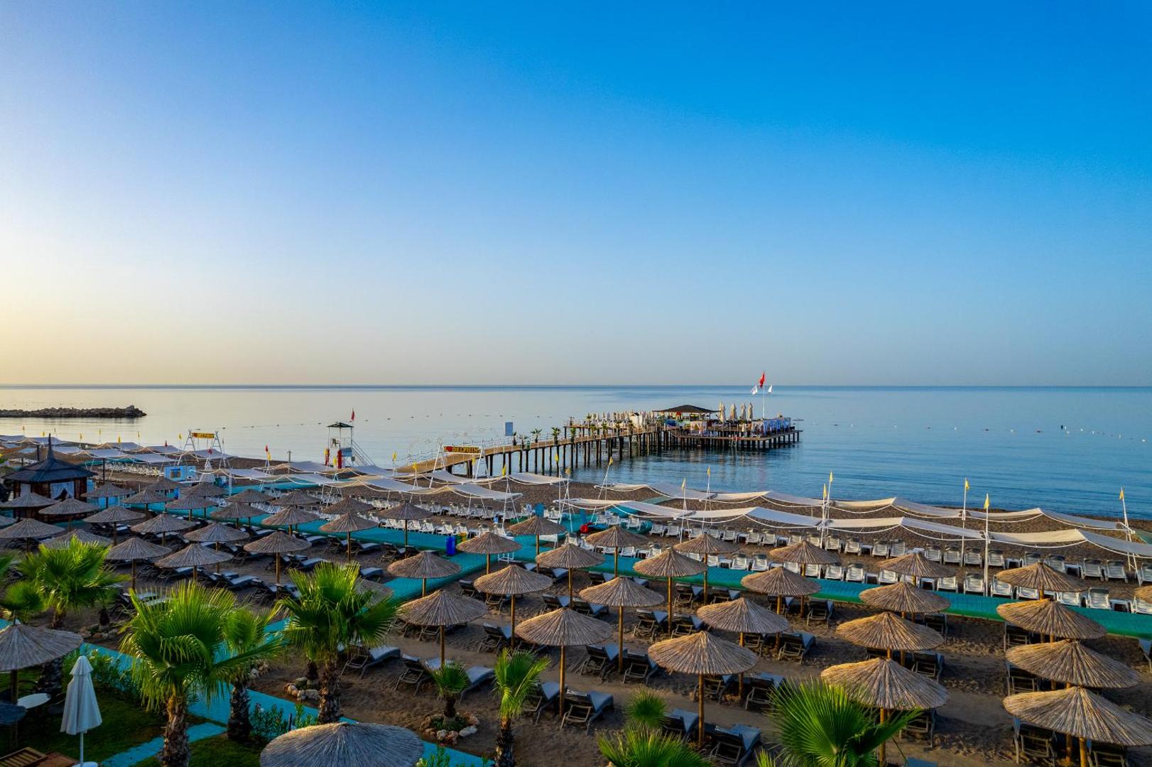 Delphin Be Grand Resort privātā pludmale ar saulessargiem, sauļošanās krēsliem un garu koka piestātni jūrā
