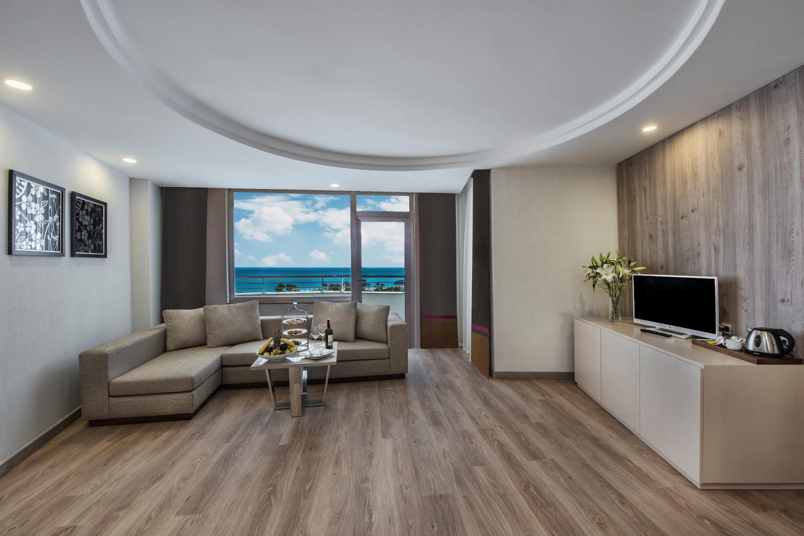 Delphin Be Grand Resort viesistabas zona ar stūra dīvānu un skatu uz Vidusjūru caur lielu logu Antalijā