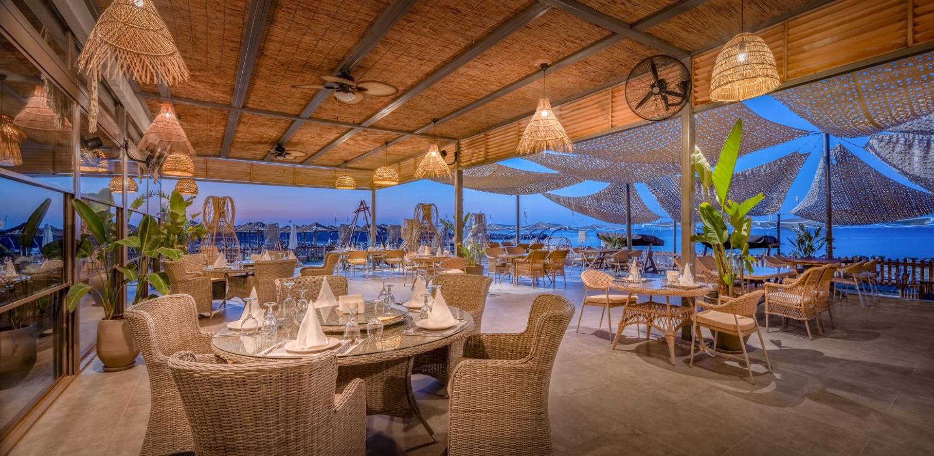 Delphin Be Grand Resort āra terases restorāns ar pītiem krēsliem un skatu uz jūru saulrietā