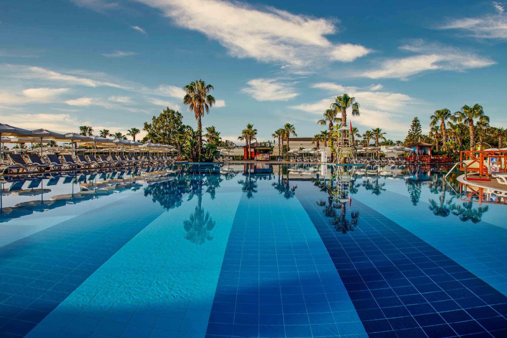 Delphin Be Grand Resort lielais āra baseins ar dzidru ūdeni, palmām un sauļošanās krēsliem zem skaidrām debesīm
