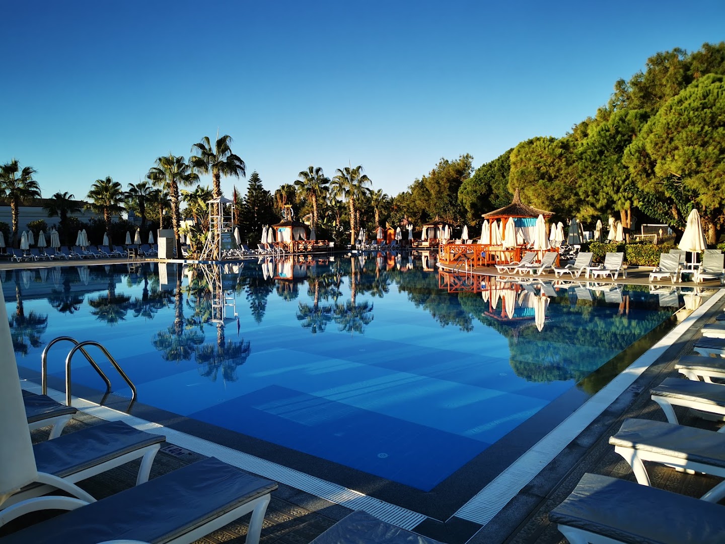 Vakara noskaņa pie Delphin Be Grand Resort baseina — koka terase ar sauļošanās krēsliem un palmām atspulgu ūdenī