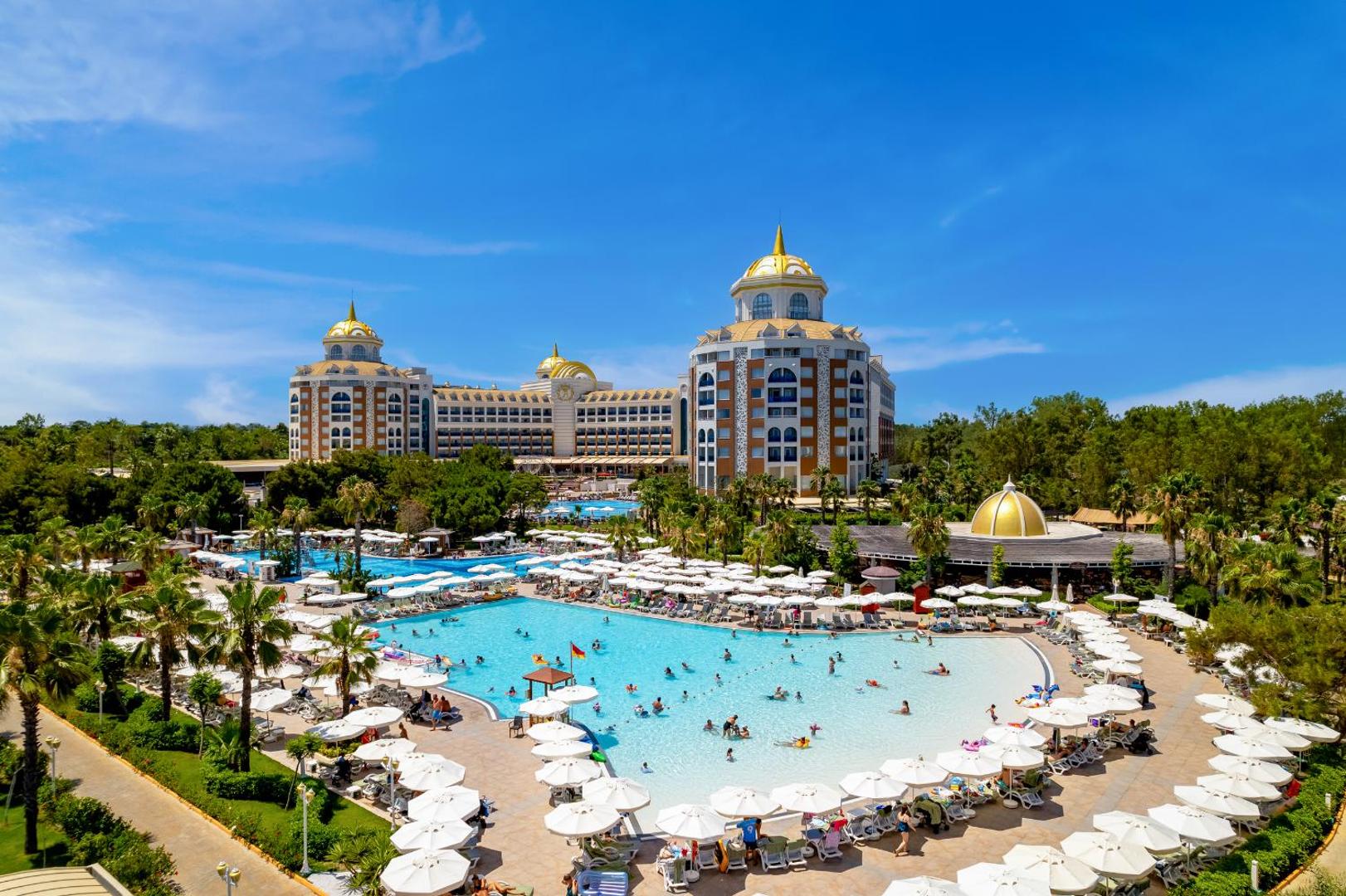Delphin Be Grand Resort panorāmas skats uz galveno baseinu un viesnīcas ēkām — plaša teritorija ar dārziem un atpūtas vietām