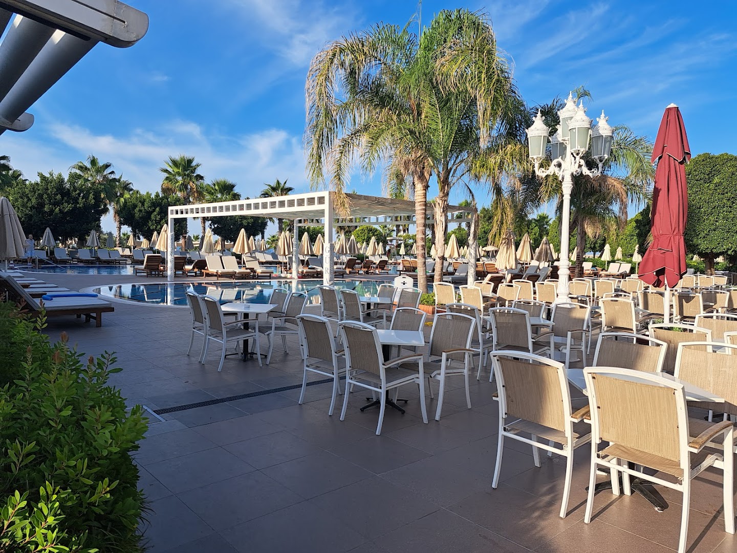 Gural Premier Belek āra terase ar galdiņiem pie baseina un palmām — atpūtas zona ar saulessargiem un ērtiem krēsliem dienas atpūtai