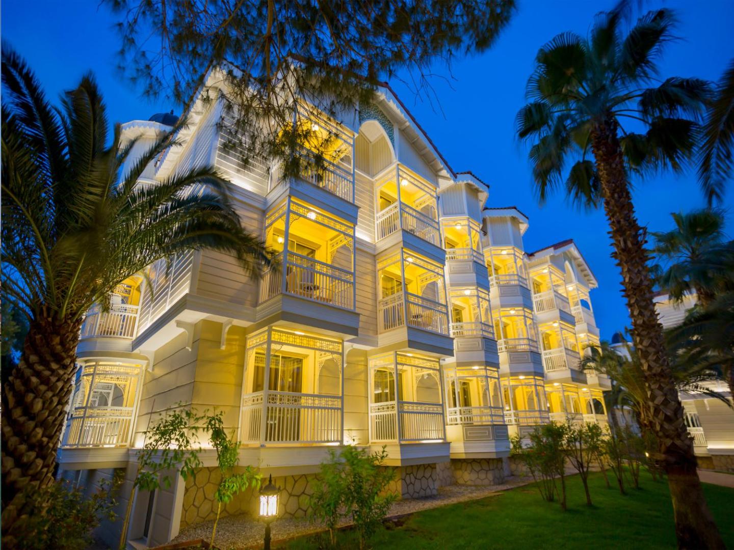 Gural Premier Belek viesnīcas ēkas fasāde vakarā ar izgaismotiem balkoniem un palmām Belekas reģionā, Turcijā