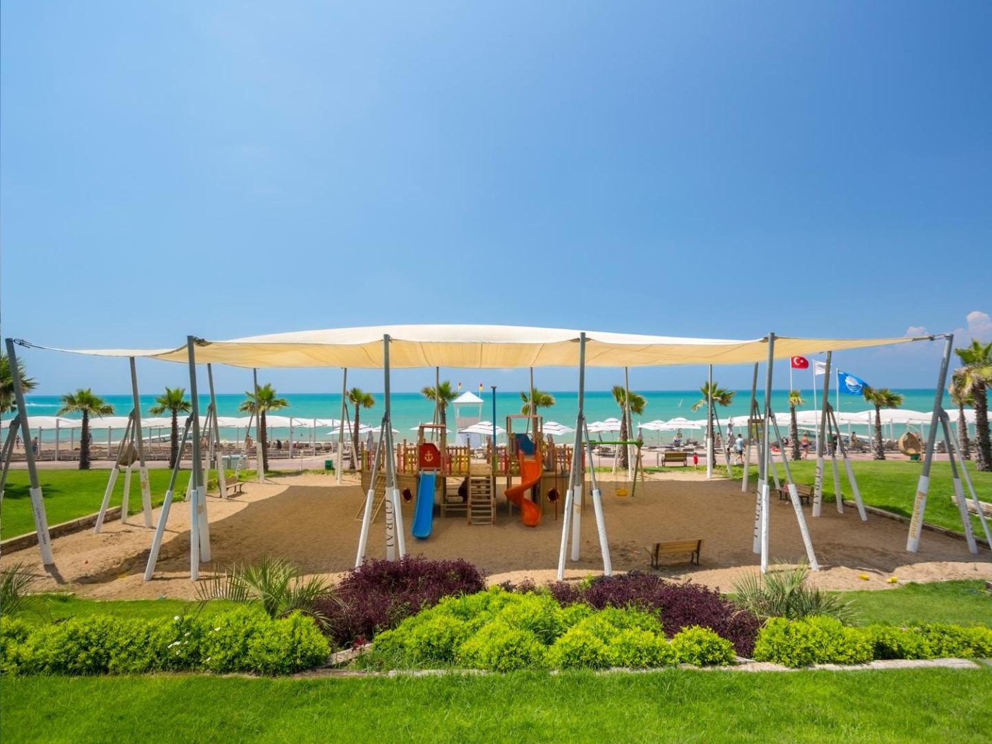 Gural Premier Belek bērnu rotaļu laukums smiltīs zem saules aizsargnojumes pie pludmales un zaļa zālāja