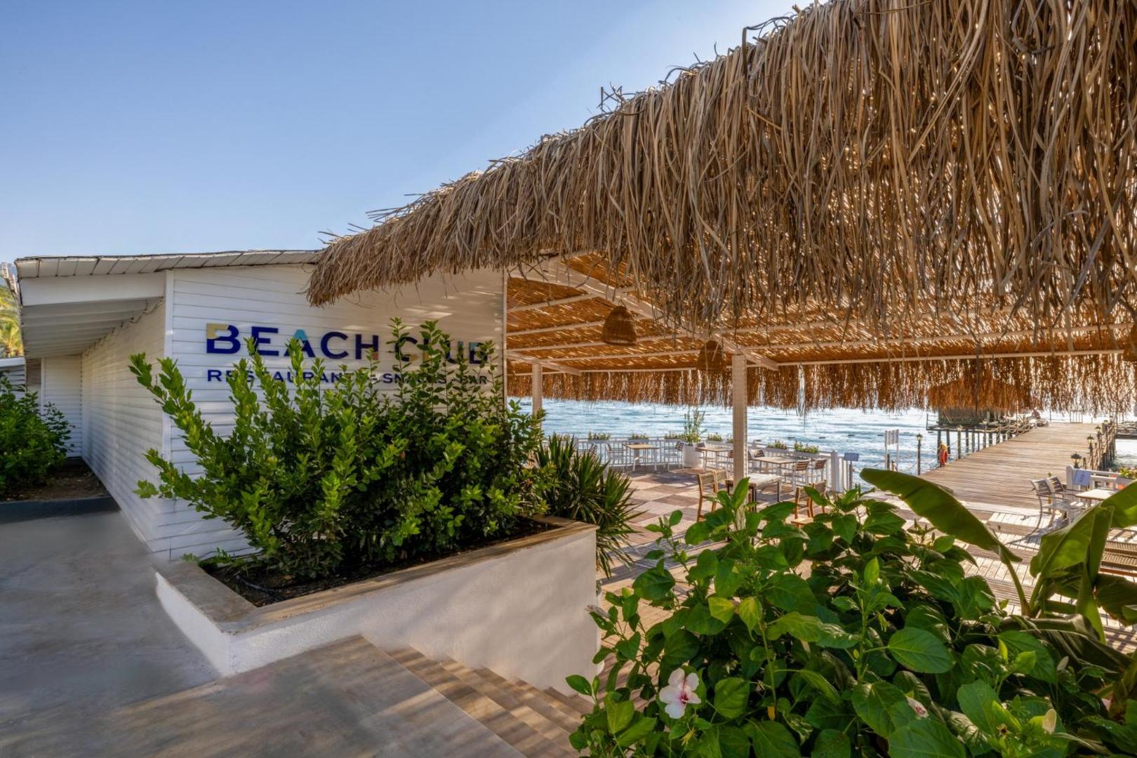 Crystal Flora Beach Resort pludmales klubs Beach Club ar salmu jumtu un ieeju uz koka piestātni pie jūras