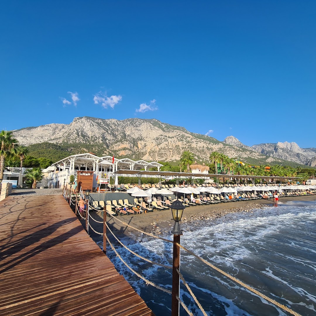 Skats no Crystal Flora Beach Resort koka laipas uz pludmali, saulessargiem un majestātiskajiem kalniem fonā saulainā dienā.