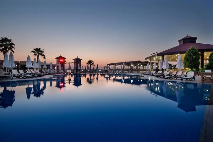 Crystal Flora Beach Resort āra baseins saulrieta laikā Beldibi reģionā — izgaismota atpūtas zona ar saulessargiem un mierīgu ūdens virsmu