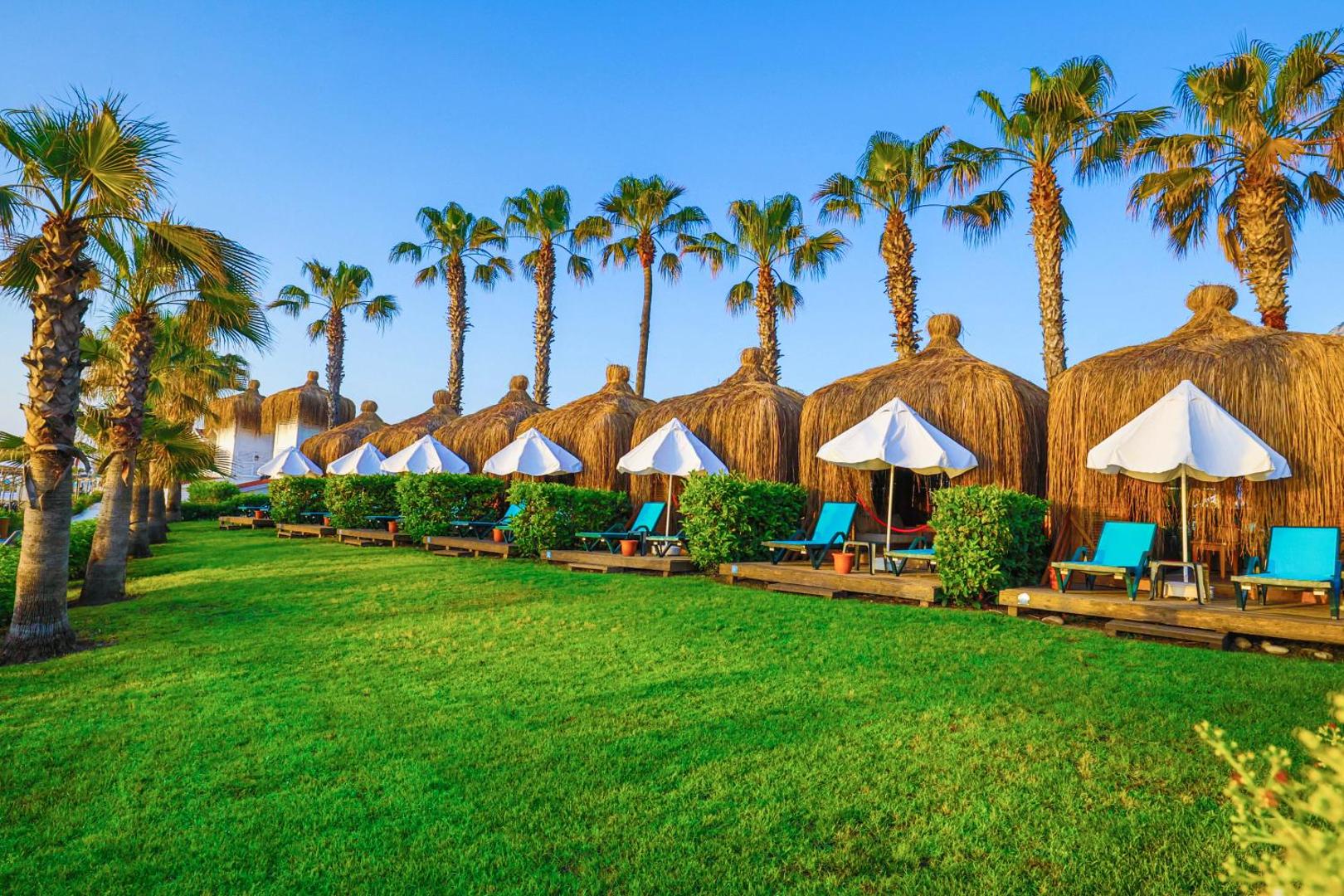 Crystal Flora Beach Resort atpūtas zona ar privātām bungalo stila nojumēm zālienā, ko ieskauj palmas un balti saulessargi Kemeras piekrastē.