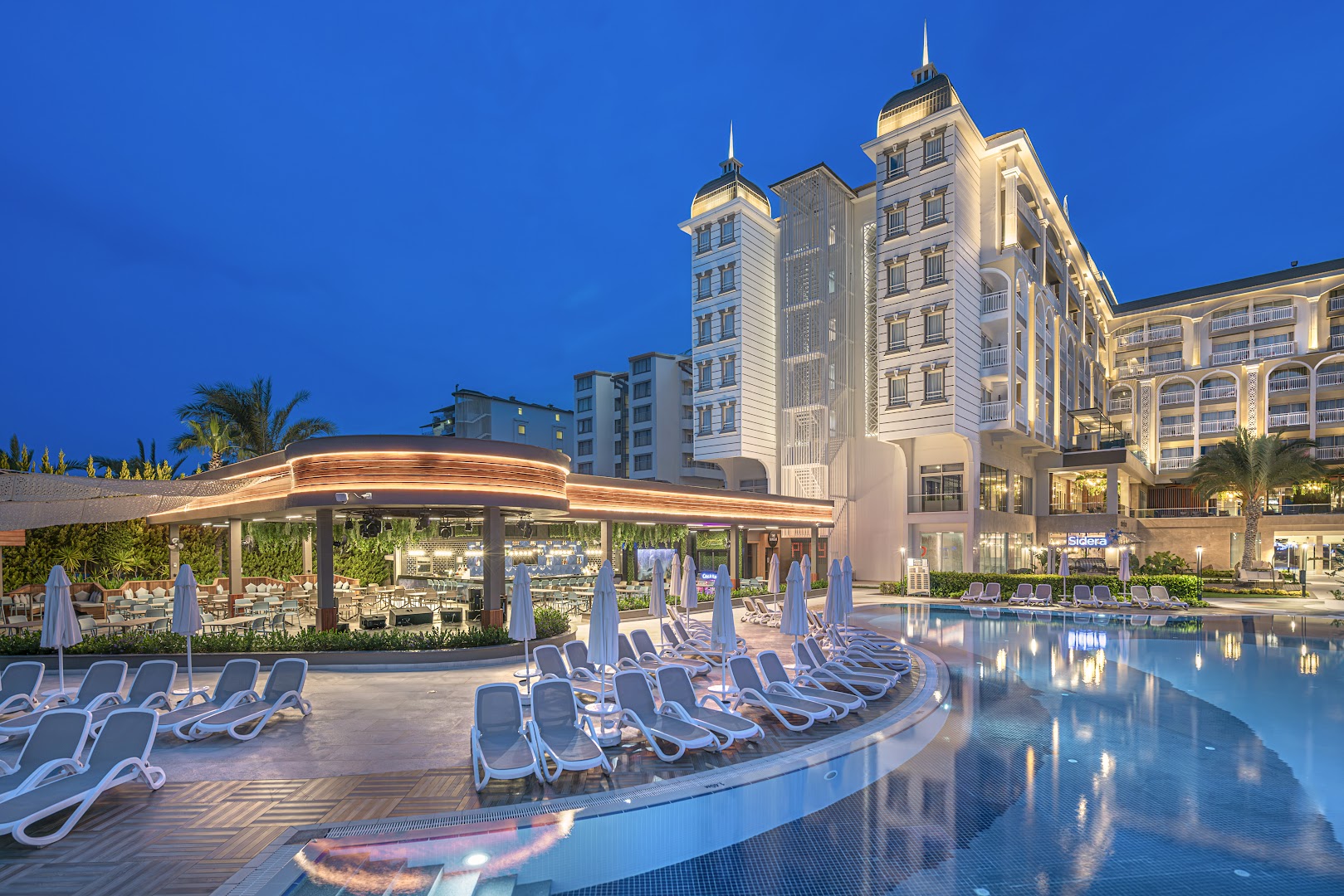Kirman Sidera Luxury & Spa viesnīcas galvenā ēka un āra baseins vakarā Sides reģionā, Turcijā — izgaismota fasāde un atpūtas zona