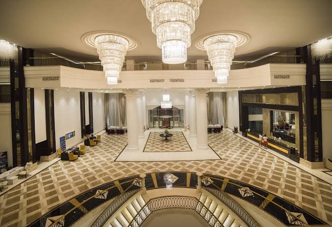 Juju Premier Palace viesnīcas lobby ar iespaidīgām kristāla lustrām, plašām kāpnēm un greznu marmora grīdas rakstu centrālajā hallē.