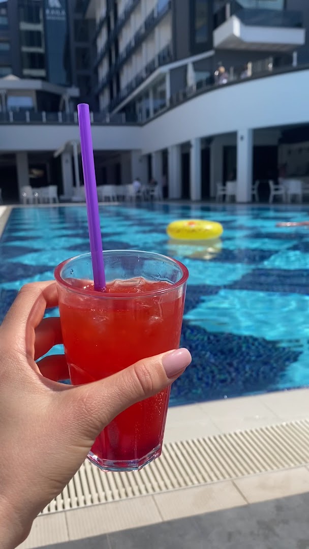 Atsvaidzinošs sarkans kokteilis pie Kaila Beach Hotel baseina — fonā redzams dzidrs ūdens un viesnīcas korpuss Alanjā