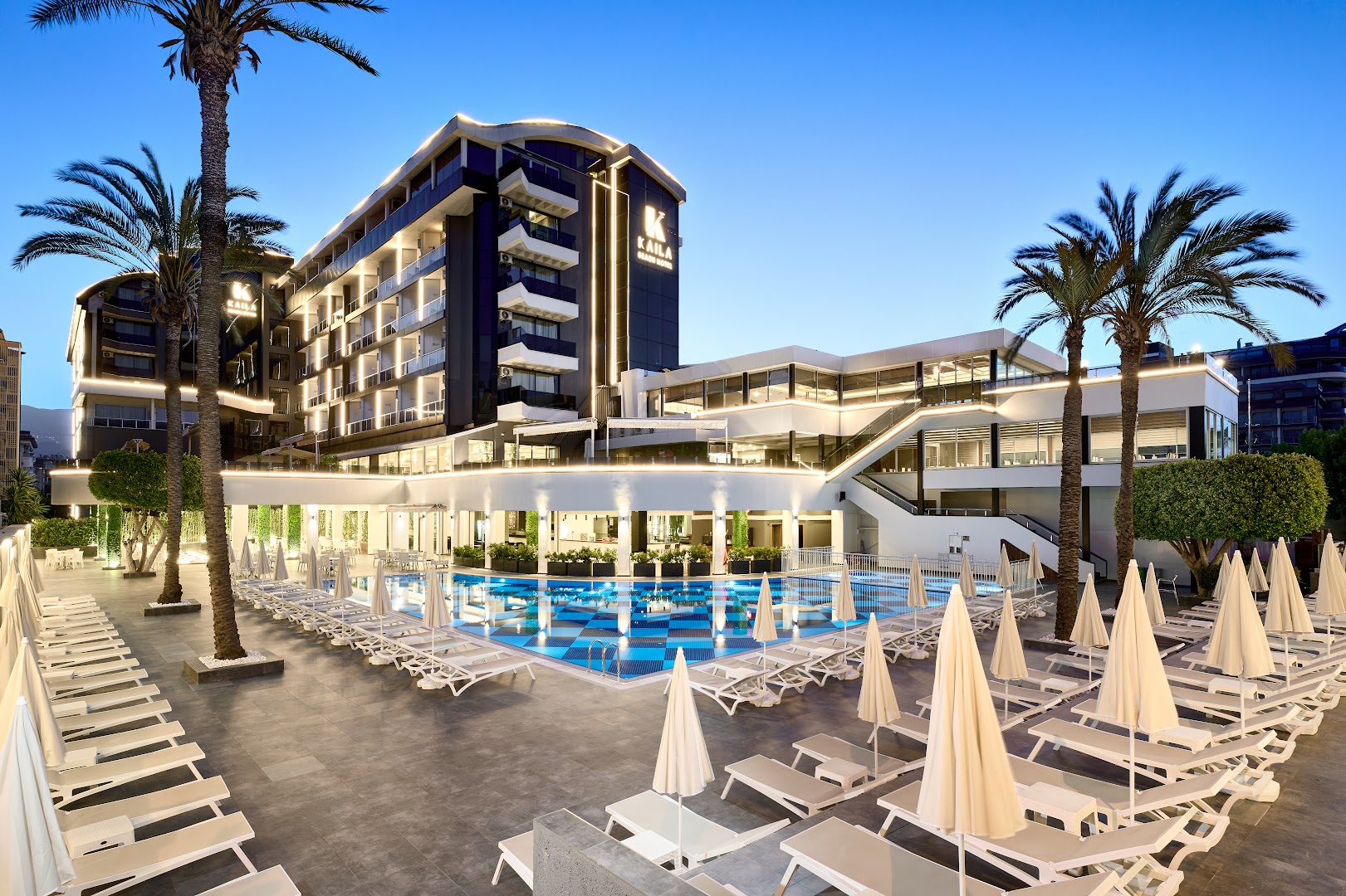 Kaila Beach Hotel galvenā ēka un izgaismots āra baseins vakarā Alānijā — moderna arhitektūra ar palmām un plašu terasi