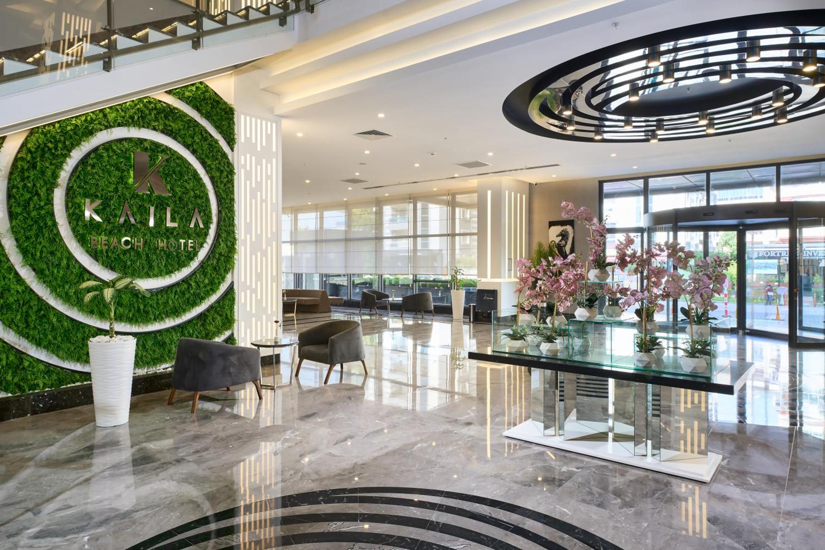 Kaila Beach Hotel plašais vestibils ar marmora grīdām, dekoratīvu zaļo sienu ar viesnīcas logo un atpūtas zonu pie ieejas