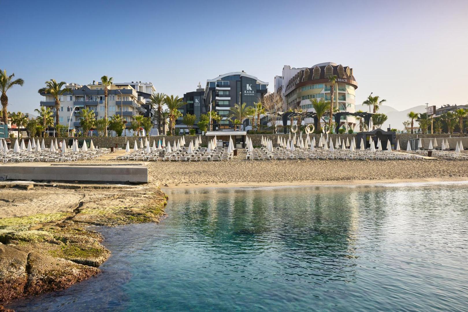 Kaila Beach Hotel skats no jūras puses uz smilšu pludmali ar saulessargiem un viesnīcas ēkām Alanjas piekrastē, Turcijā